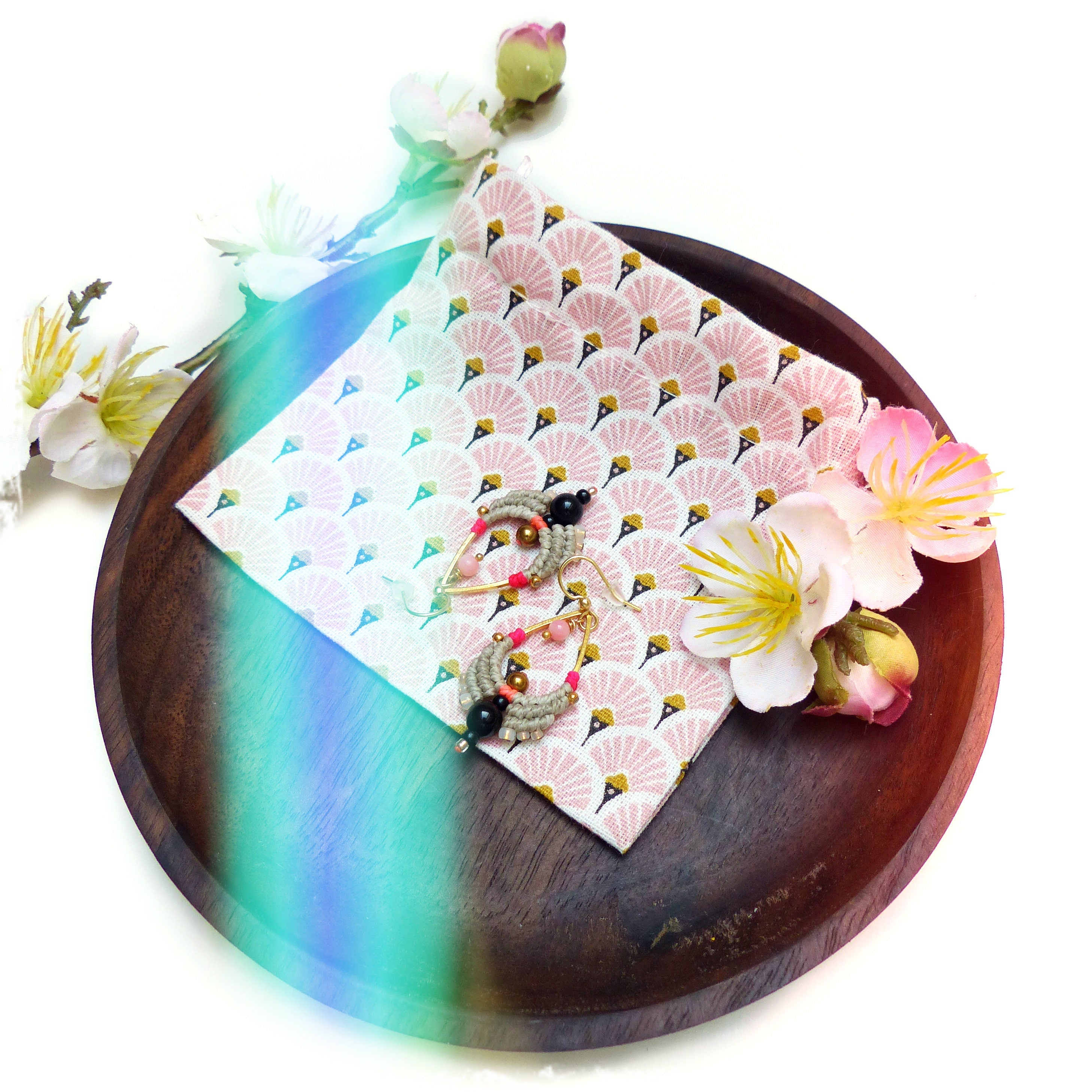 Peut inclure: Une paire de boucles d'oreilles faites à la main avec des perles noires, brunes et roses sur une base de fil doré. Les boucles d'oreilles sont présentées sur un tissu à motifs roses et blancs avec un motif floral. Le tissu est sur un plateau en bois.