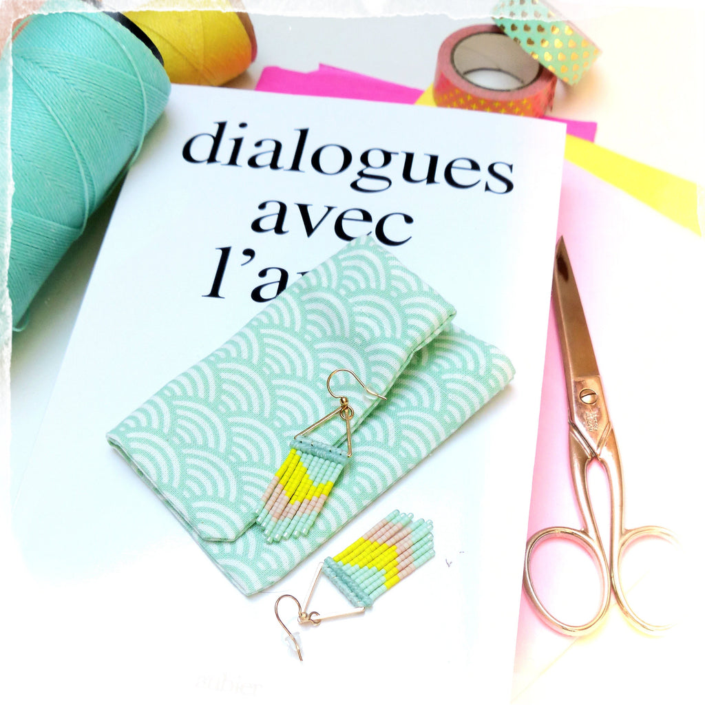 Peut inclure: Une paire de boucles d'oreilles en franges de perles turquoise et jaunes avec des crochets dorés. Les boucles d'oreilles sont présentées sur une petite pochette en tissu à motif vert et blanc. La pochette est sur une surface blanche avec une paire de ciseaux et un livre avec le texte "dialogues avec l'a" visible en arrière-plan.
