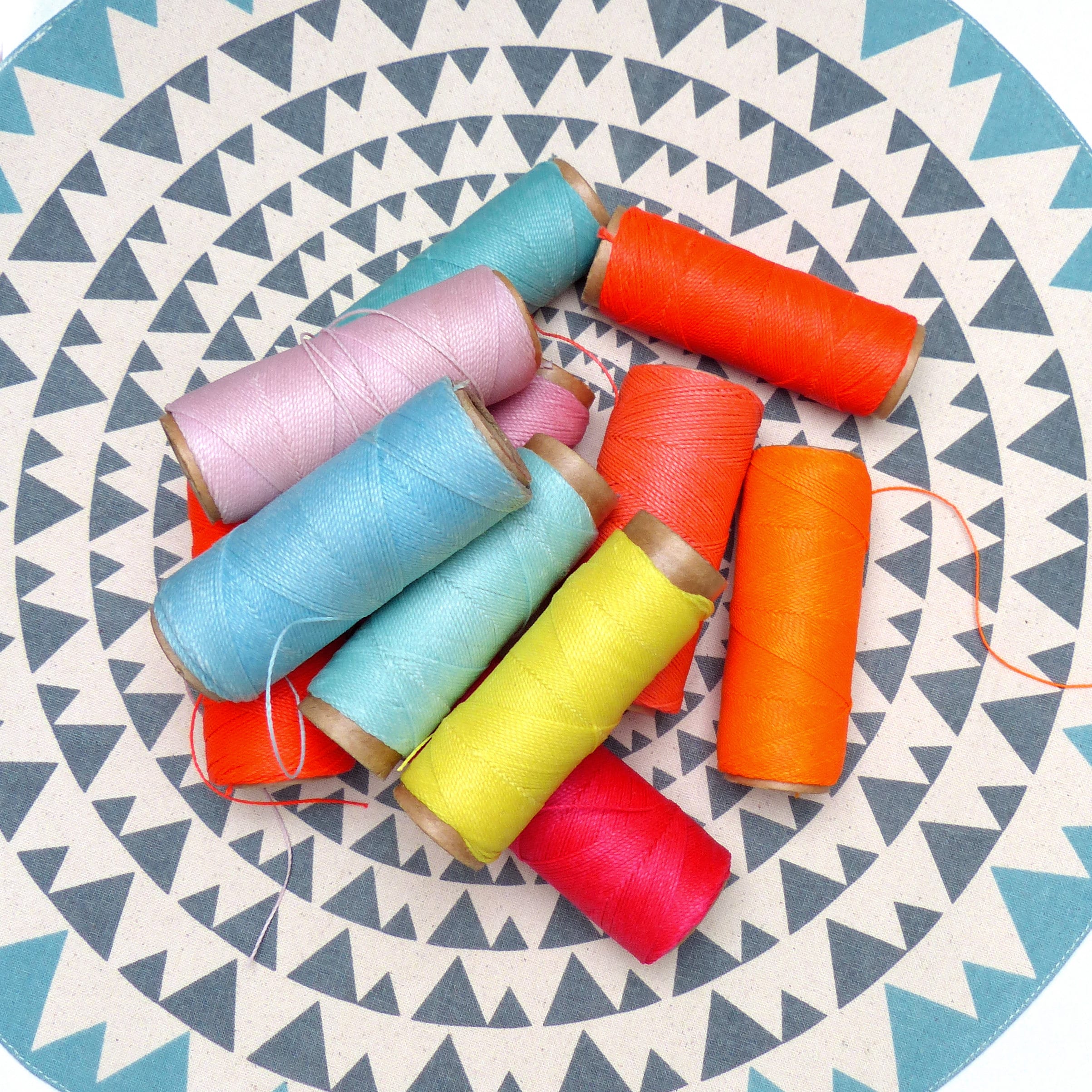 Peut inclure: Une collection de bobines de fil colorées dans différentes nuances de rose, bleu, jaune, orange et rouge. Les bobines sont disposées en cercle sur une surface à motifs blancs et gris.