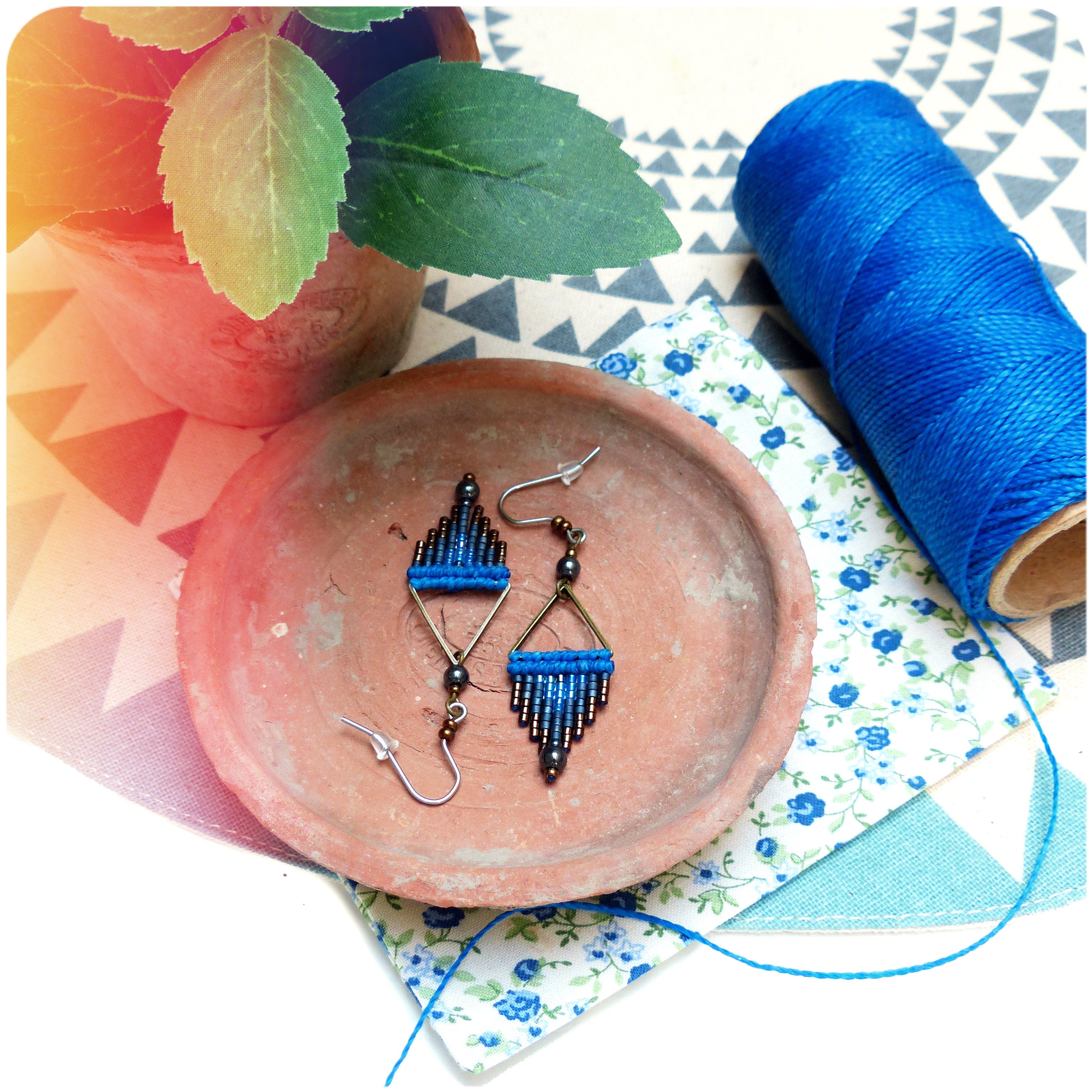 Peut inclure: Une paire de boucles d'oreilles géométriques bleues et brunes en forme de triangle. Les boucles d'oreilles sont sur une assiette en terre cuite avec un motif floral bleu et blanc.