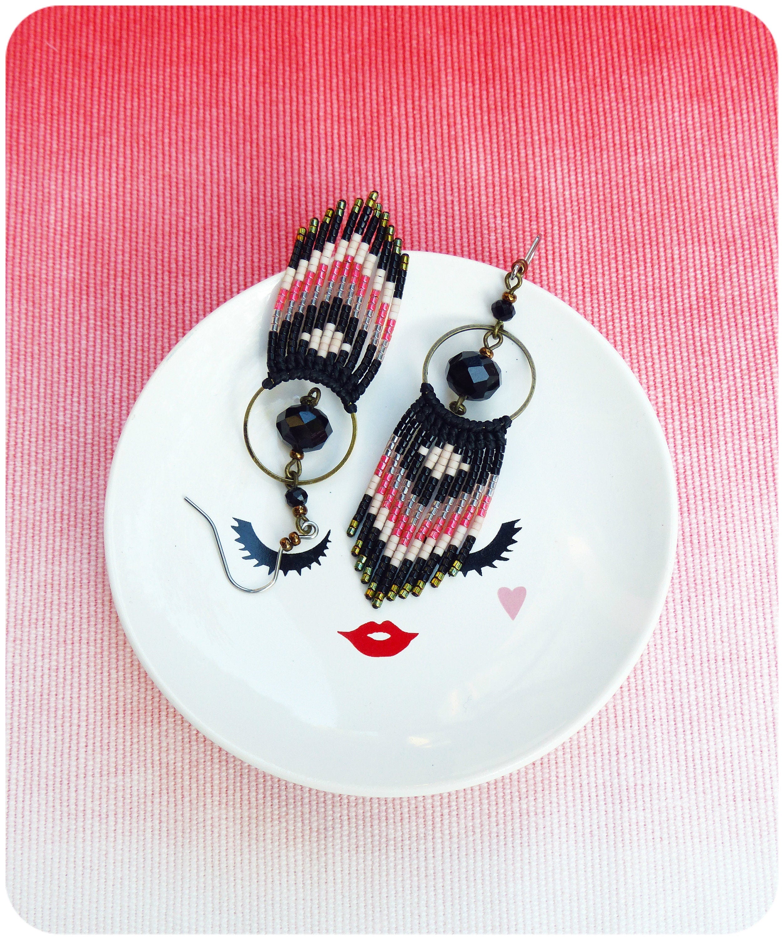 Peut inclure: Une paire de boucles d'oreilles en perles noires, blanches et roses avec des breloques circulaires noires suspendues à une assiette blanche avec un visage stylisé.
