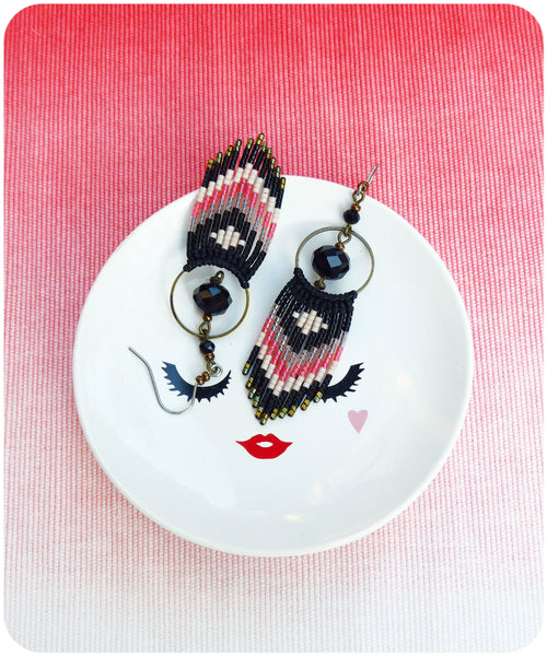 Peut inclure: Une paire de boucles d'oreilles en perles noires, blanches et roses avec des breloques circulaires noires suspendues à une assiette blanche avec un visage stylisé.