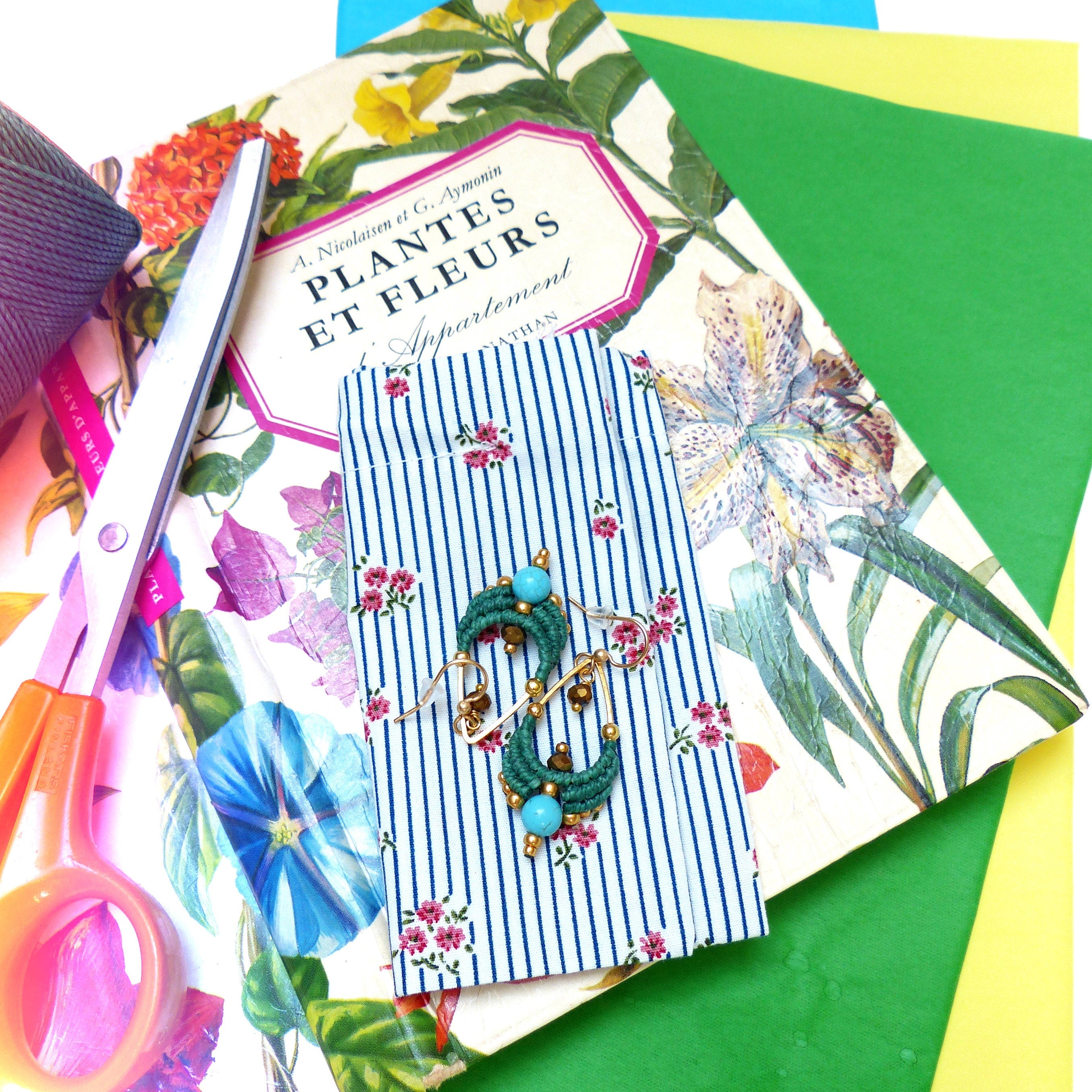 Peut inclure: Une paire de boucles d'oreilles turquoise et dorées avec un motif floral sur une pochette en tissu rayée bleu et blanc. La pochette est posée sur un livre intitulé "Plantes et Fleurs" en français.