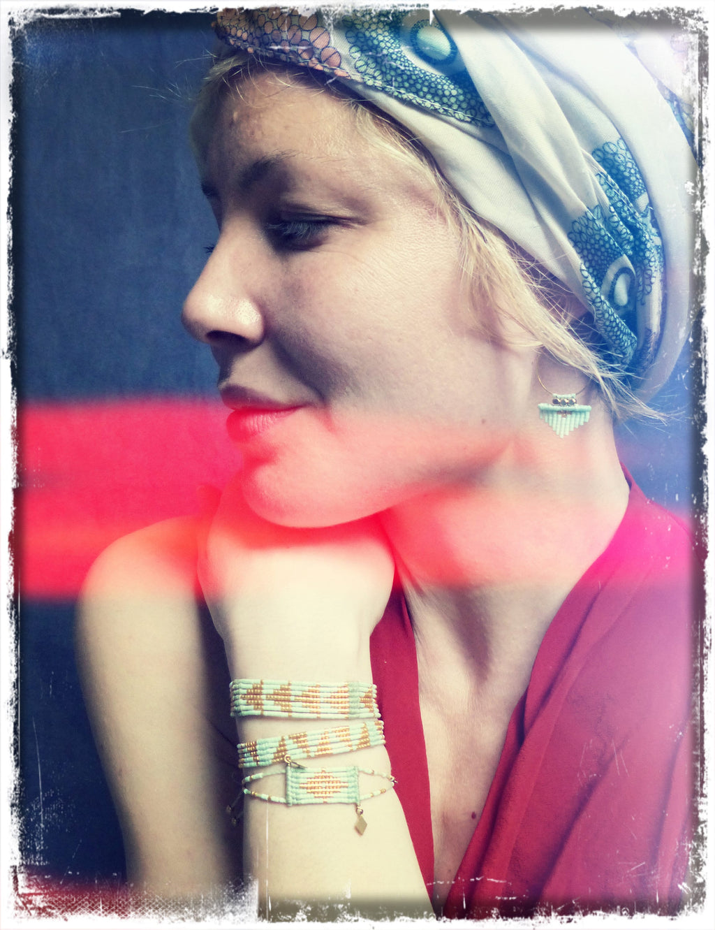 Peut inclure: Une femme portant un foulard blanc à motif floral, un haut rouge et plusieurs bracelets en perles au poignet. La femme regarde sur le côté et a le menton posé sur sa main.