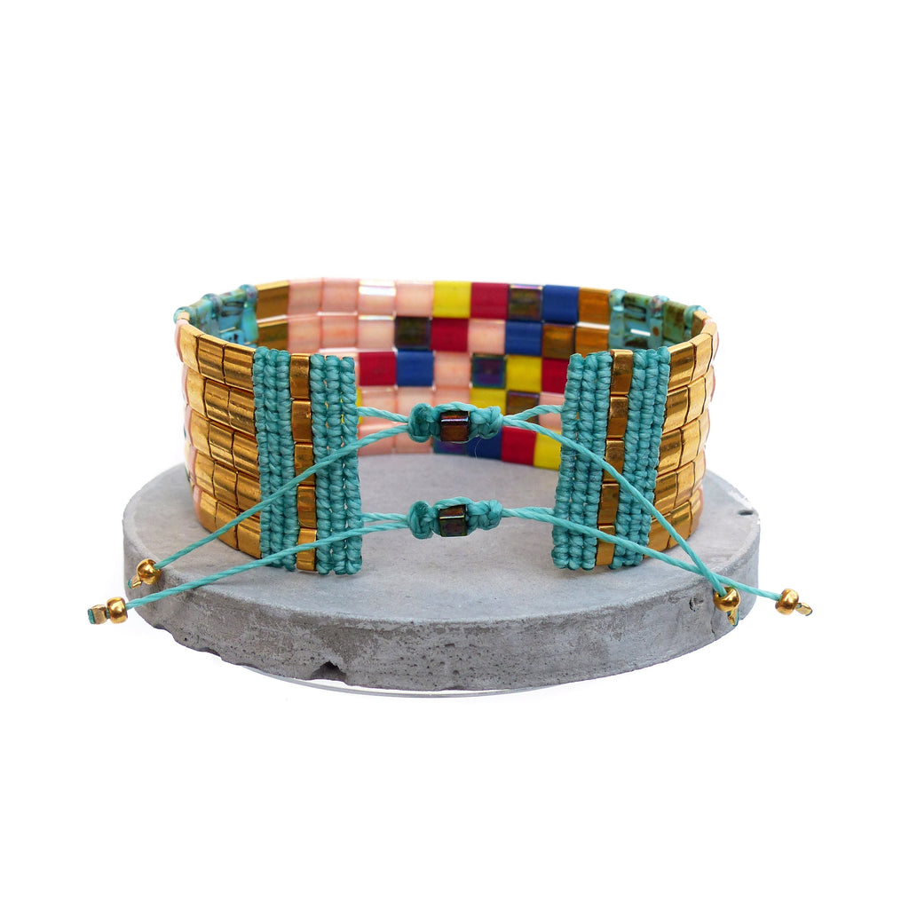 Peut inclure: Un bracelet de perles avec un design doré, turquoise et multicolore. Le bracelet présente des perles rectangulaires dorées, des accents turquoise et un mélange de perles carrées roses, jaunes, bleues et rouges. Il est présenté sur une surface grise et circulaire.