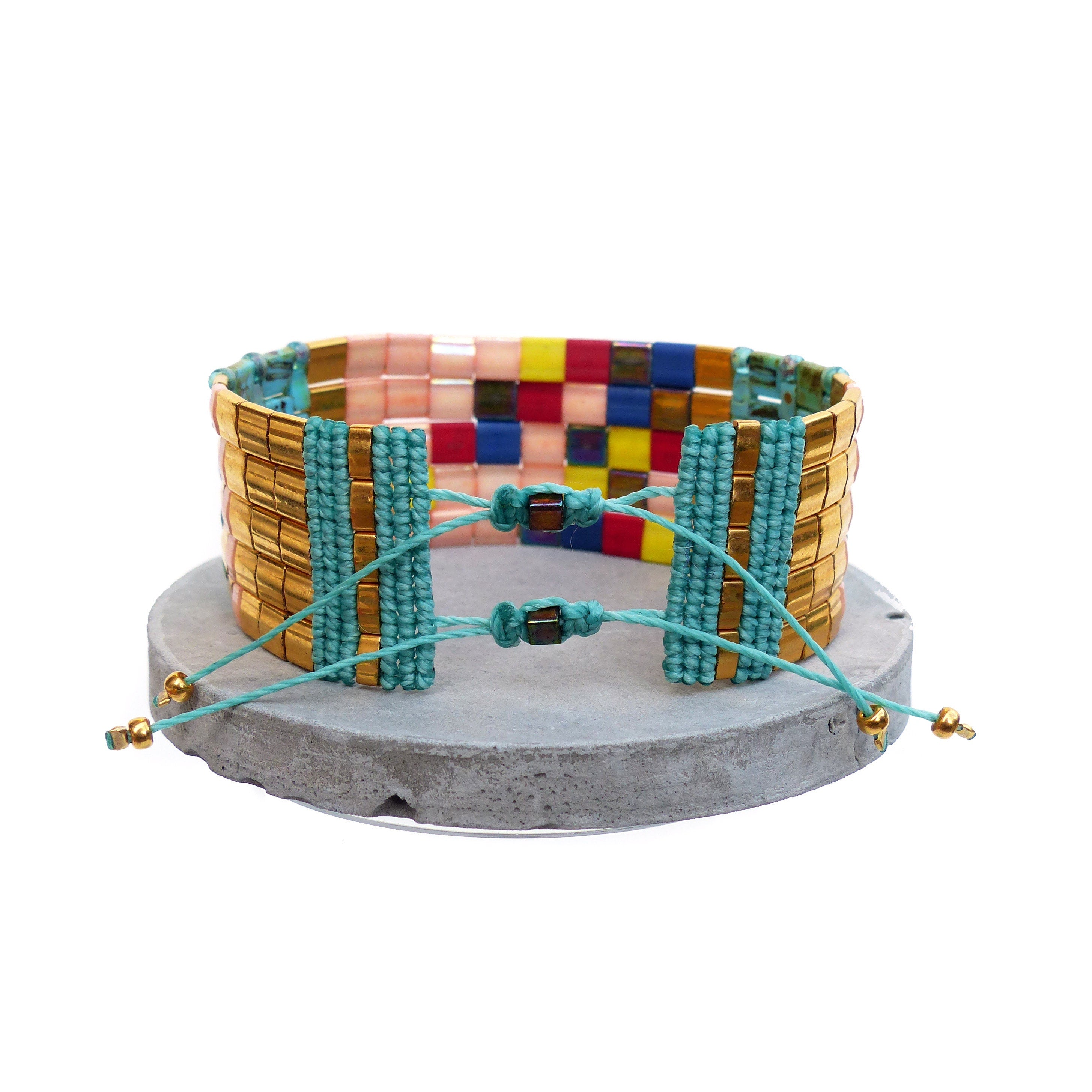 Peut inclure: Un bracelet de perles avec un design doré, turquoise et multicolore. Le bracelet présente des perles rectangulaires dorées, des accents turquoise et un mélange de perles carrées roses, jaunes, bleues et rouges. Il est présenté sur une surface grise et circulaire.