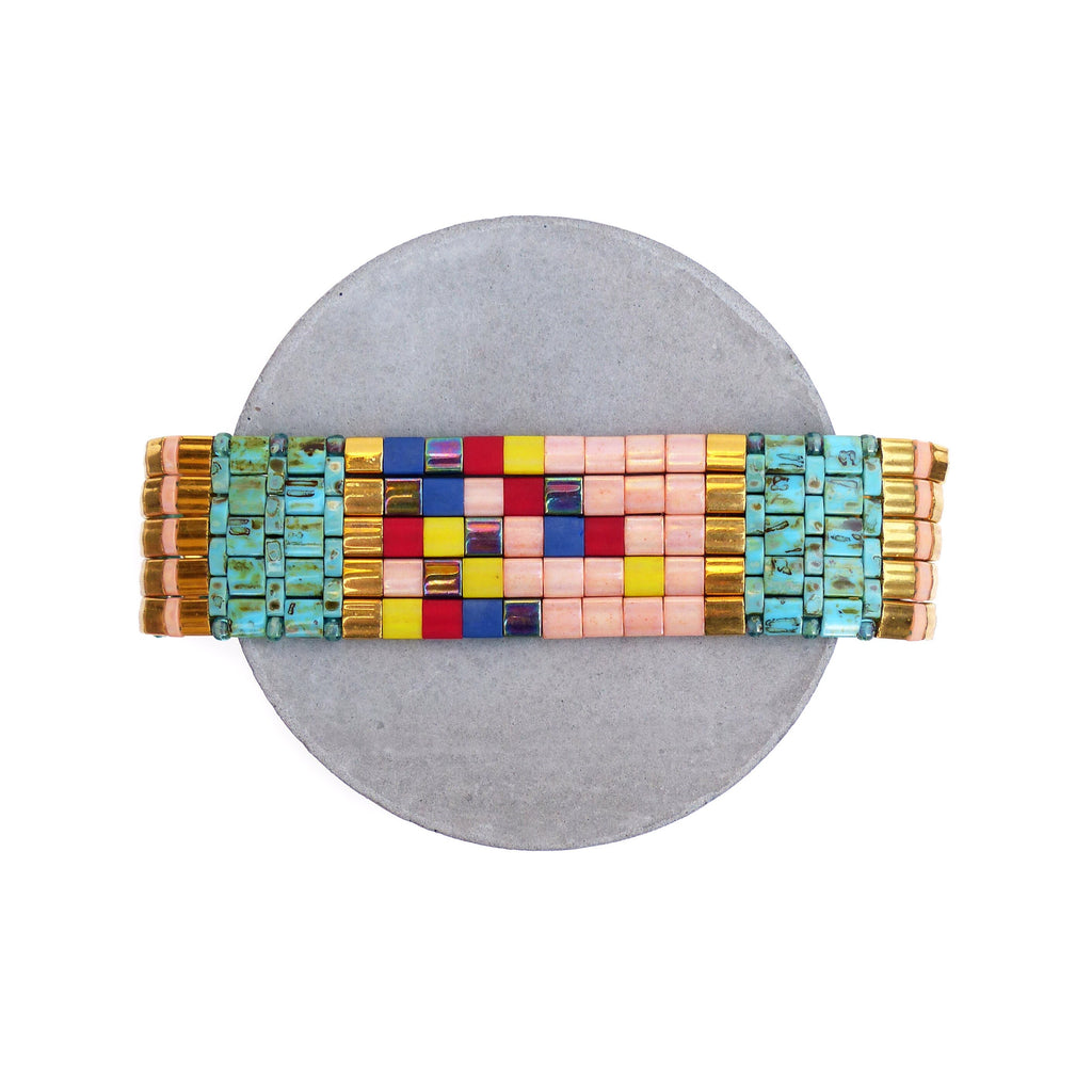 Peut inclure: Un bracelet de perles colorées avec un motif géométrique. Le bracelet présente un mélange de perles roses, dorées, turquoise, jaunes, rouges et bleues. Il est présenté sur une surface grise et circulaire sur fond blanc.