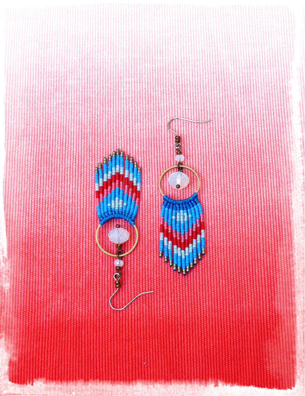 Peut inclure: Une paire de boucles d'oreilles en perles bleues, rouges et blanches avec un charme en cristal et un anneau en or. Les boucles d'oreilles sont suspendues à un fond rouge.