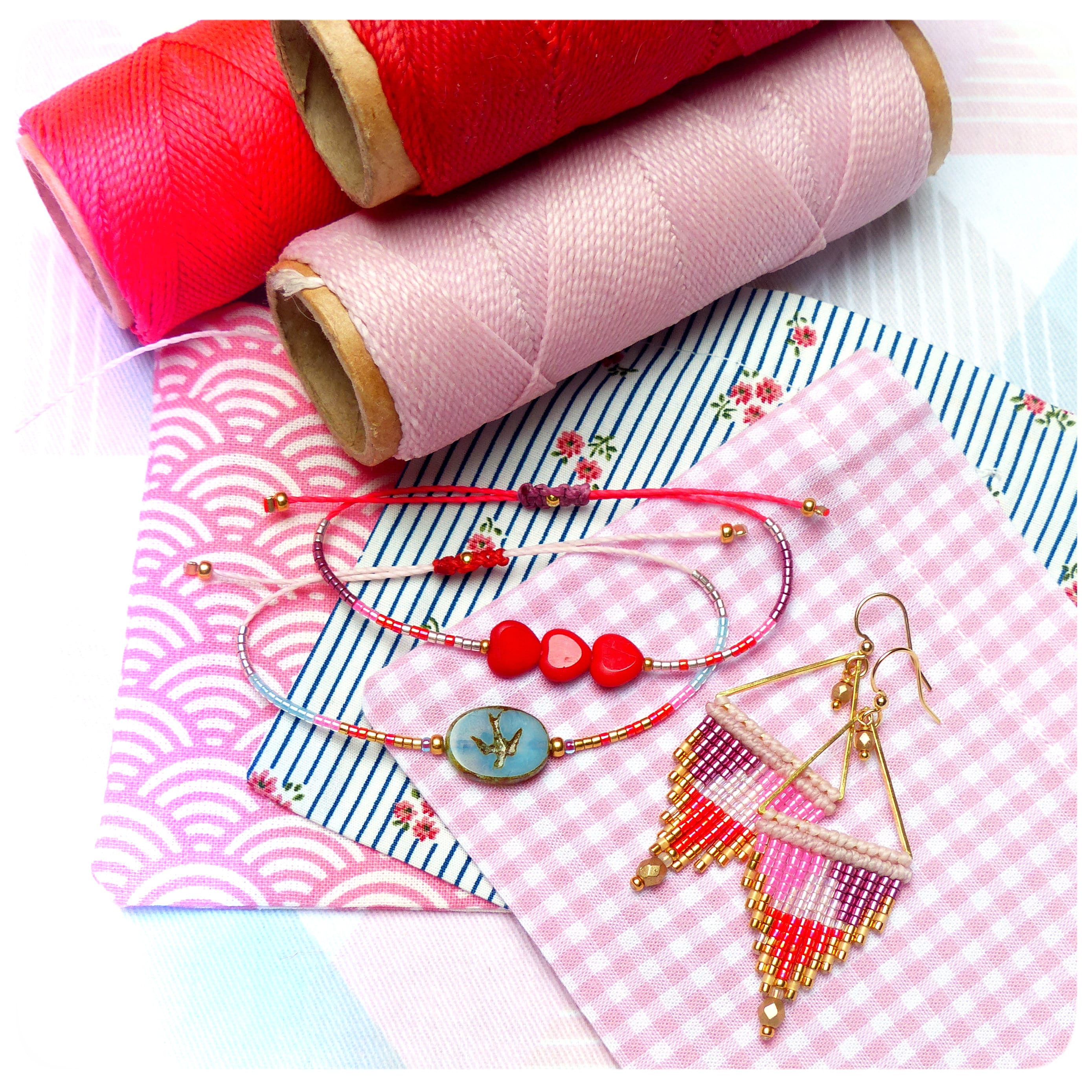 Peut inclure: Deux bracelets roses et rouges avec des perles dorées et un charme d'oiseau bleu, et une paire de boucles d'oreilles dorées avec des perles roses, rouges et blanches. Les bijoux sont sur un tissu à carreaux rose et blanc.