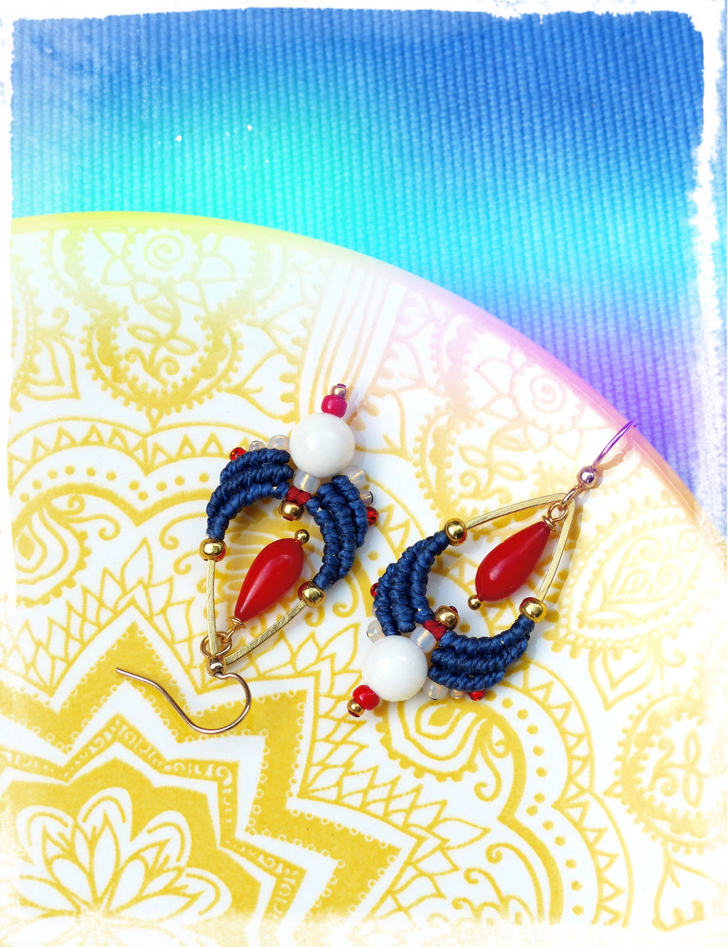 Peut inclure: Une paire de boucles d'oreilles faites à la main avec du macramé bleu et blanc, des perles rouges et blanches et des accents dorés. Les boucles d'oreilles sont présentées sur une assiette blanche avec un motif de mandala jaune et or.