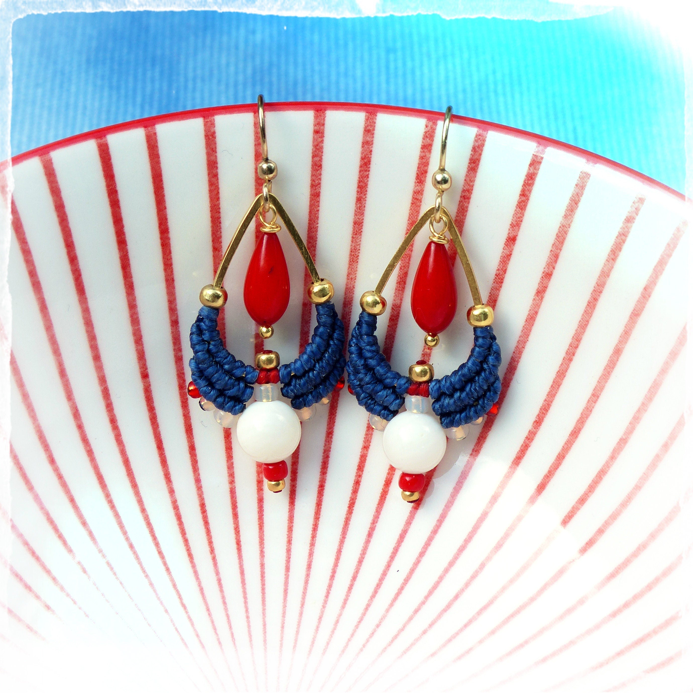 Peut inclure: Une paire de boucles d'oreilles pendantes avec un motif rouge, blanc et bleu. Les boucles d'oreilles présentent un pendentif en forme de larme de couleur dorée avec une perle rouge, un motif macramé bleu et blanc et une perle blanche.