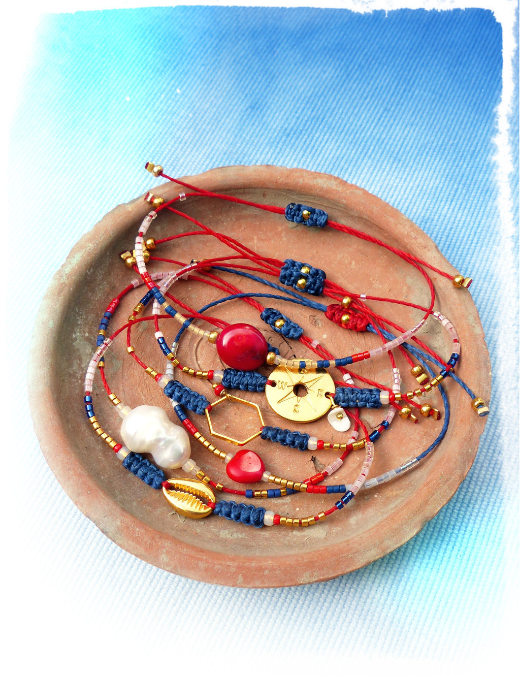 Peut inclure: Un ensemble de bracelets d'amitié rouges et bleus avec des perles et des breloques dorées, dont une boussole, un coquillage et un hexagone. Les bracelets sont présentés sur une assiette en terre cuite.