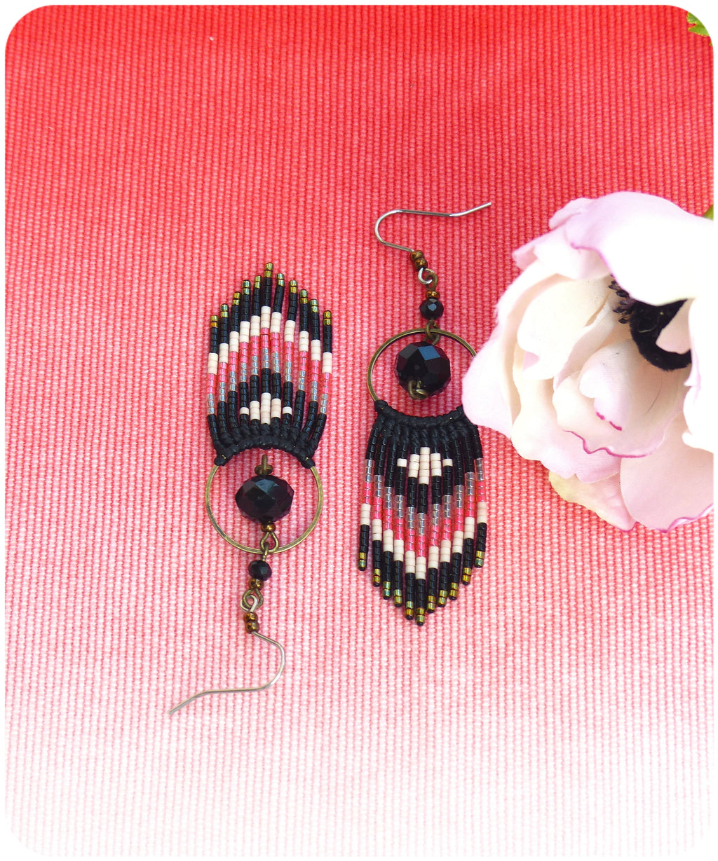 Peut inclure: Une paire de boucles d'oreilles en perles noires, blanches et roses avec une perle noire et un pendentif en forme de cercle doré. Les boucles d'oreilles sont sur un fond texturé rose.