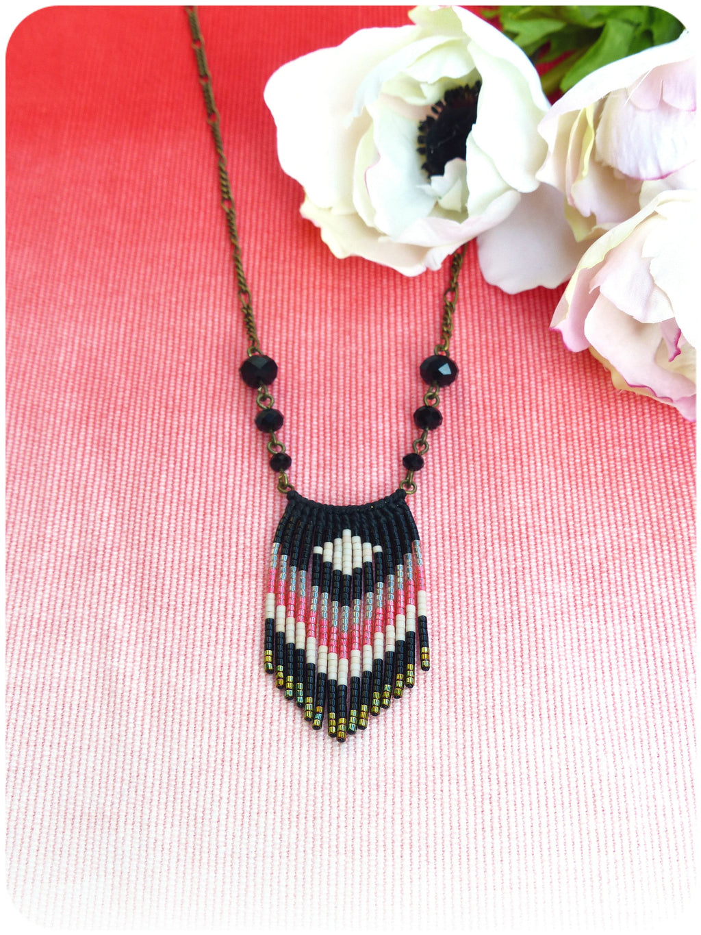 Peut inclure: Un collier de perles noires et blanches avec une frange rose et dorée. Le collier est sur un fond rose et blanc avec des fleurs blanches.