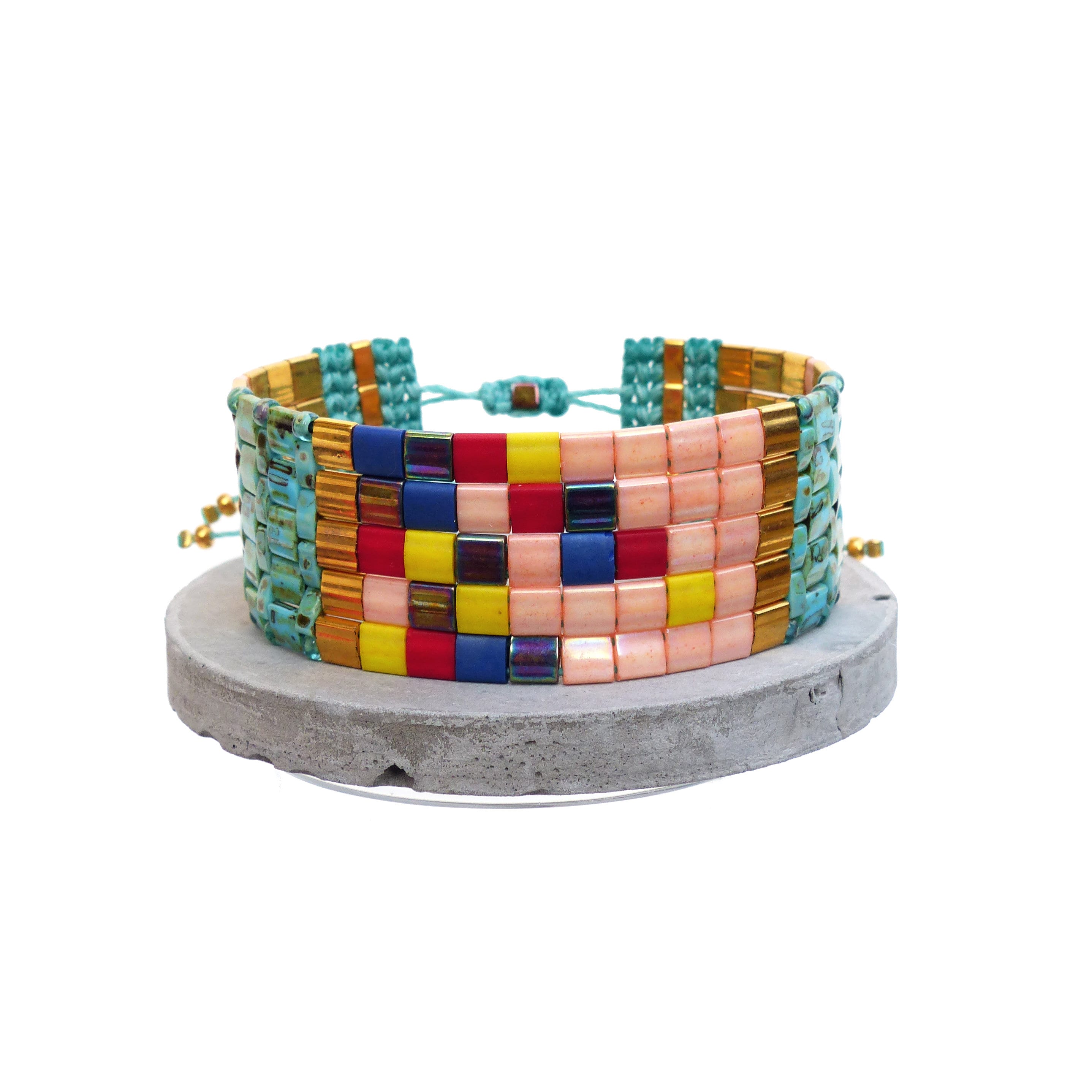 Peut inclure: Un bracelet de perles colorées avec un motif tissé. Le bracelet présente des perles carrées roses, bleues, jaunes, rouges et dorées. Le bracelet est présenté sur une surface grise et circulaire sur fond blanc.