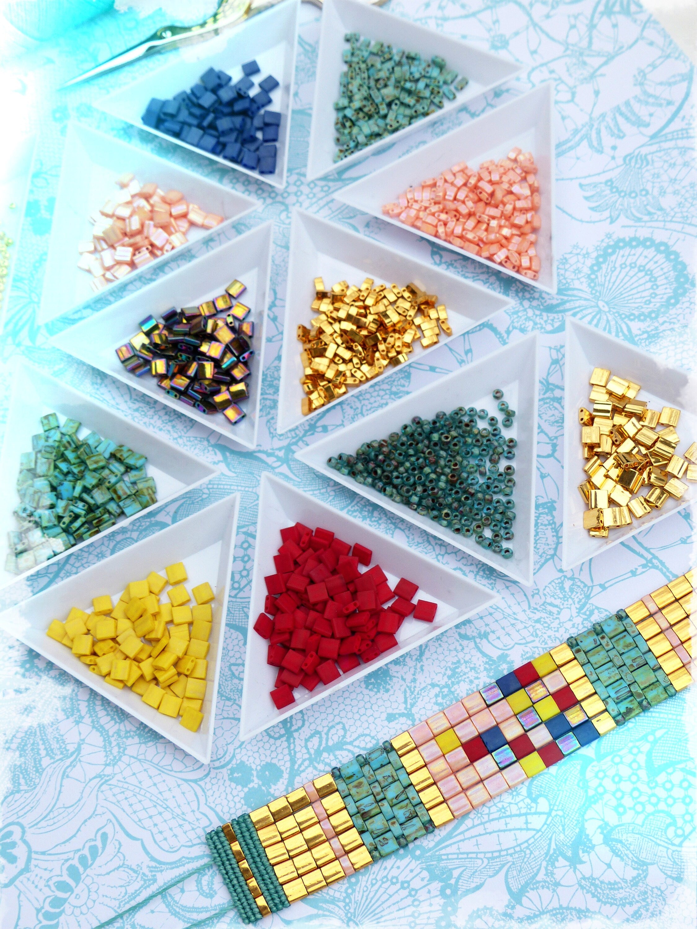 Peut inclure: Une collection de perles colorées dans des plateaux triangulaires blancs, à côté d'un bracelet de perles fini. Les perles sont de différentes couleurs, dont le bleu, l'or, le rouge et le turquoise. Le bracelet présente un motif géométrique.