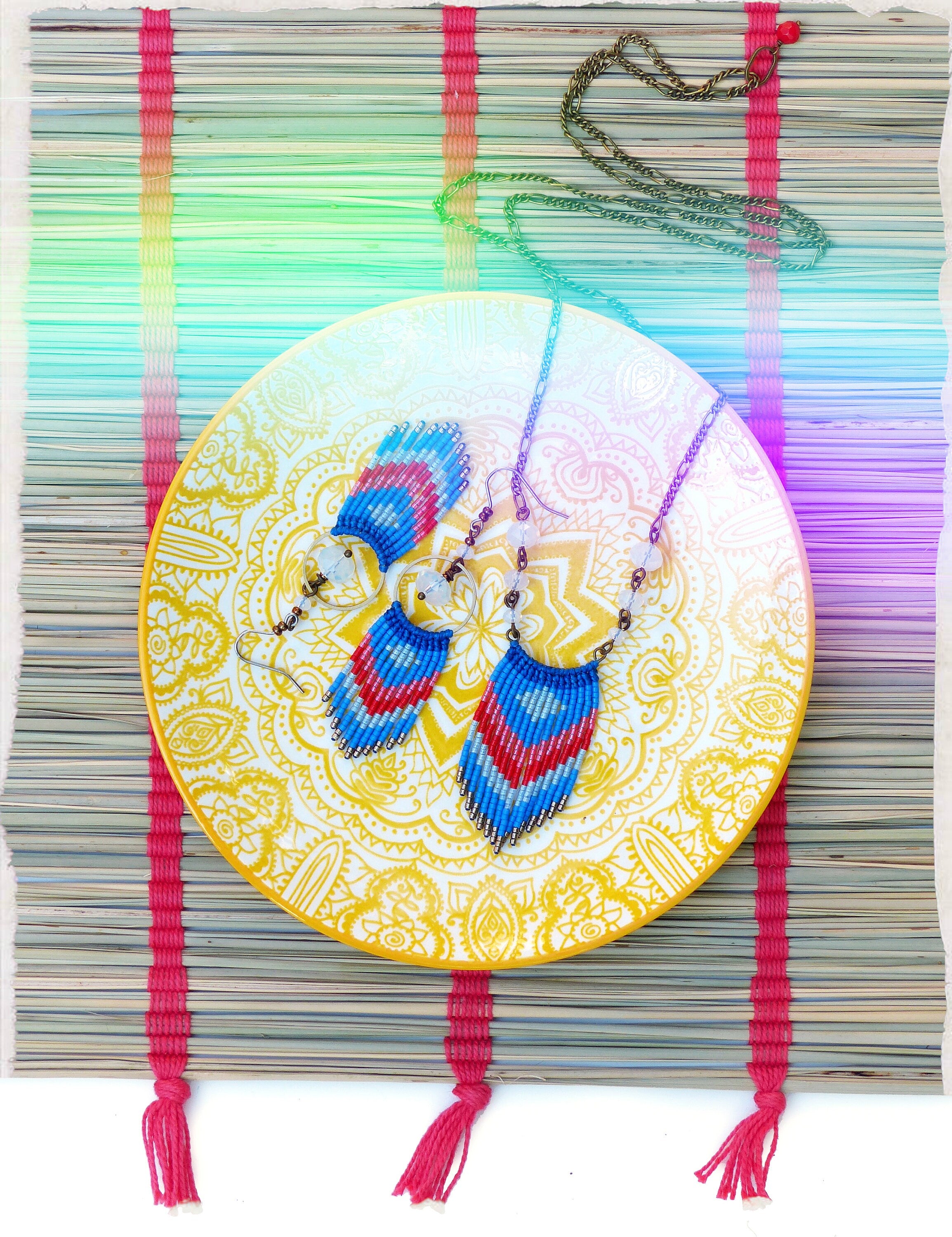 Peut inclure: Boucles d'oreilles et collier à pendentif en perles bleues, rouges et blanches, présentés sur une assiette jaune à motifs floraux blancs. L'assiette est posée sur un tapis tissé avec des détails roses.