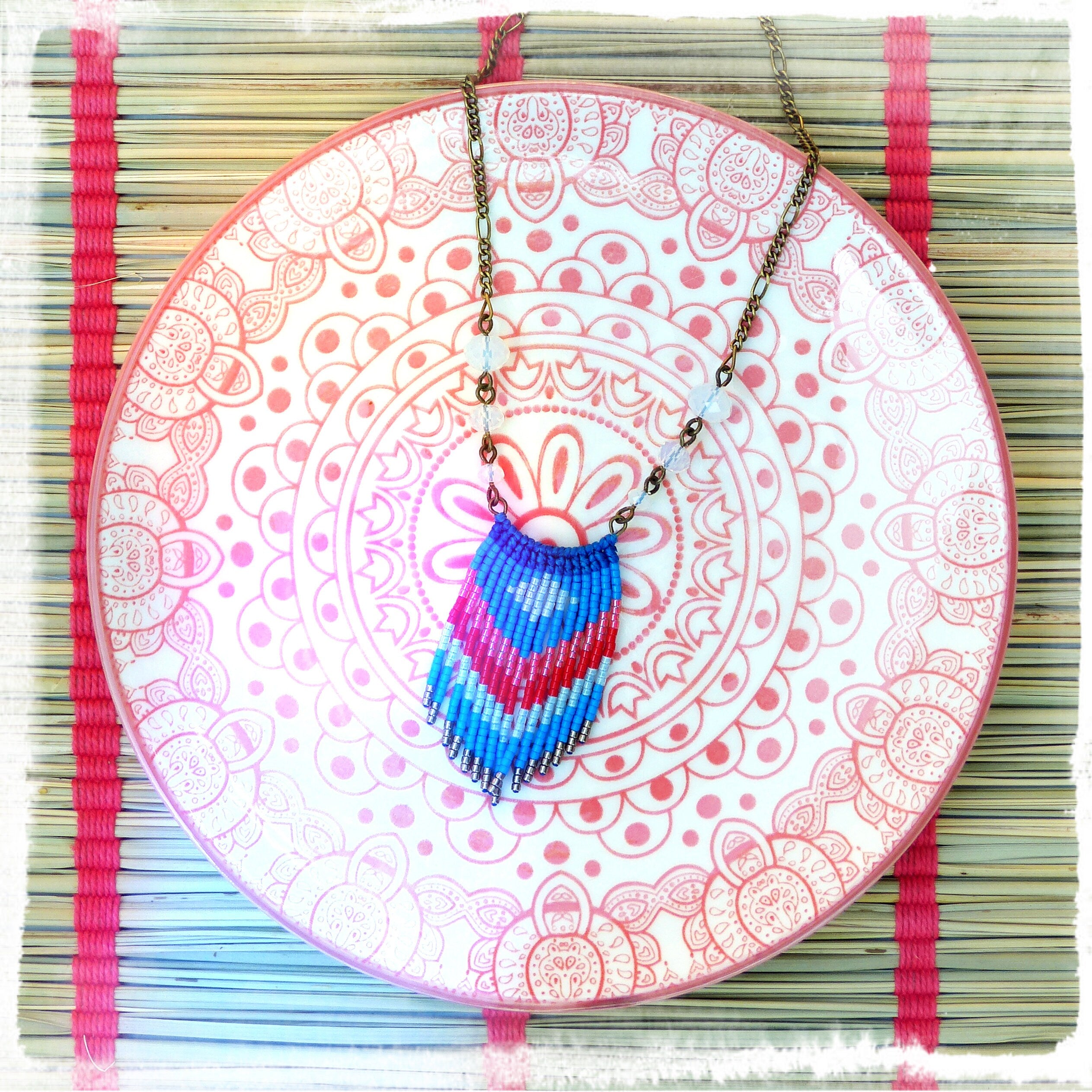 Peut inclure: Un collier en macramé coloré avec un motif géométrique bleu, rose et rouge, posé sur une assiette à motifs rose et blanc. Le collier a une chaîne en bronze et de petites perles transparentes. L'assiette repose sur un tapis en bambou.