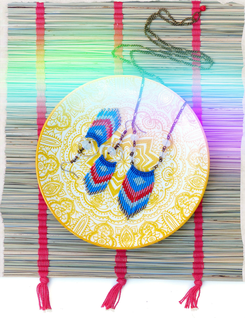 Peut inclure: Une paire de boucles d'oreilles en perles avec des rayures bleues, rouges et blanches suspendues à une assiette jaune avec un motif de mandala blanc. Les boucles d'oreilles sont suspendues à un collier à chaîne.