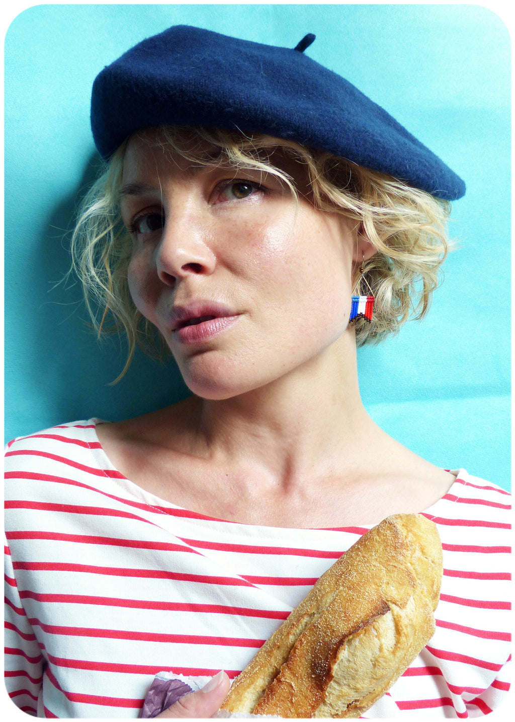 Peut inclure: Une femme portant un béret bleu et une chemise rayée rouge et blanche tient une baguette devant elle. Elle porte des boucles d'oreilles aux couleurs du drapeau français.