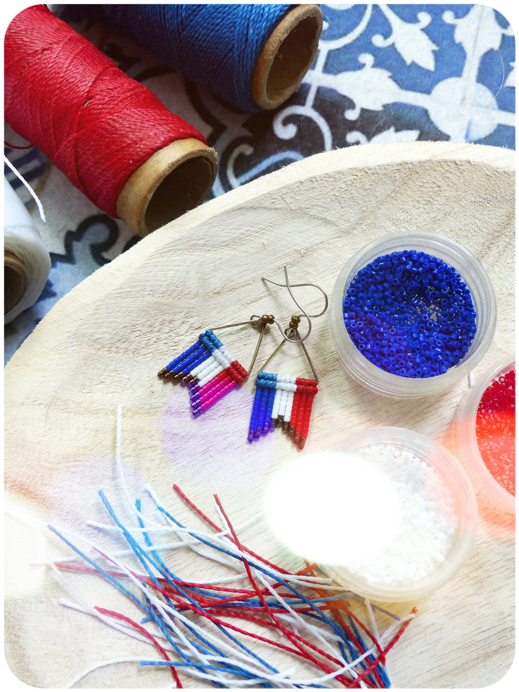 Peut inclure: Une paire de boucles d'oreilles en perles avec un motif de drapeau français, des perles rouges, blanches et bleues et un fil de couleur or. Les boucles d'oreilles sont sur une surface en bois avec des bobines de fil rouge, blanc et bleu, et de petits récipients de perles.