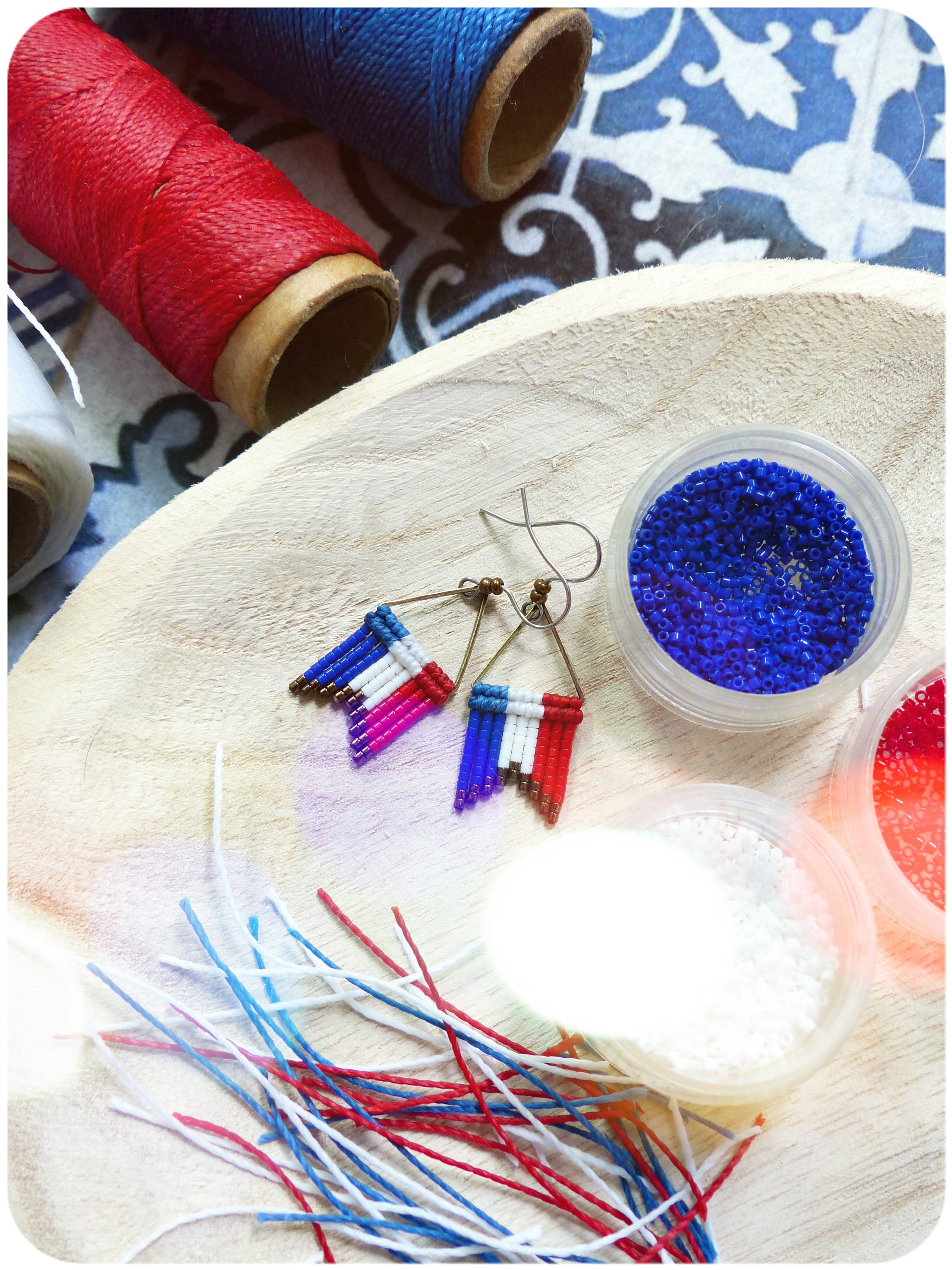 Peut inclure: Une paire de boucles d'oreilles en perles avec un motif de drapeau français, des perles rouges, blanches et bleues et un fil de couleur or. Les boucles d'oreilles sont sur une surface en bois avec des bobines de fil rouge, blanc et bleu, et de petits récipients de perles.