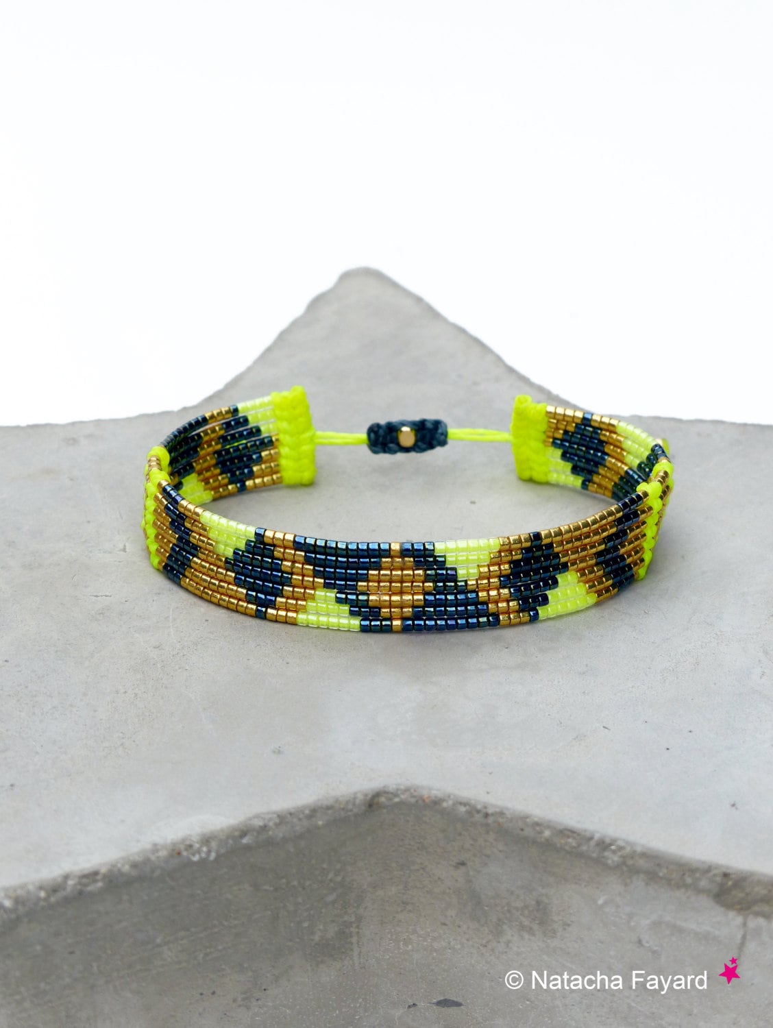 Peut inclure: Un bracelet de perles avec un motif géométrique. Le bracelet présente des perles jaune fluo, bleu marine et dorées. La fermeture réglable est un cordon bleu foncé avec une petite perle dorée. Le bracelet est présenté sur une surface grise.