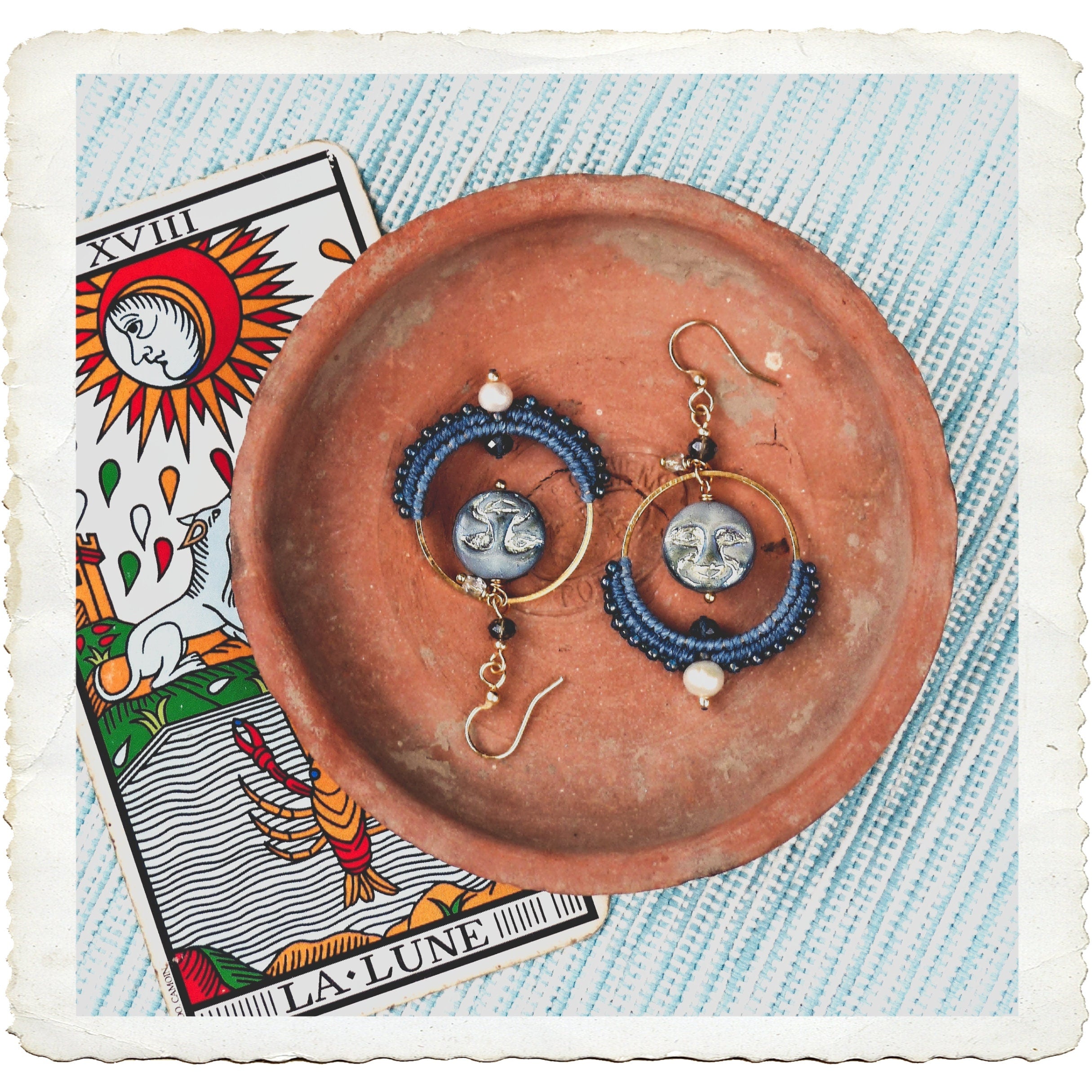 Peut inclure: Une paire de boucles d'oreilles bleues et dorées avec un charme de lune et un accent de perle blanche. Les boucles d'oreilles sont présentées sur une assiette en terre cuite avec une carte de tarot représentant la lune.