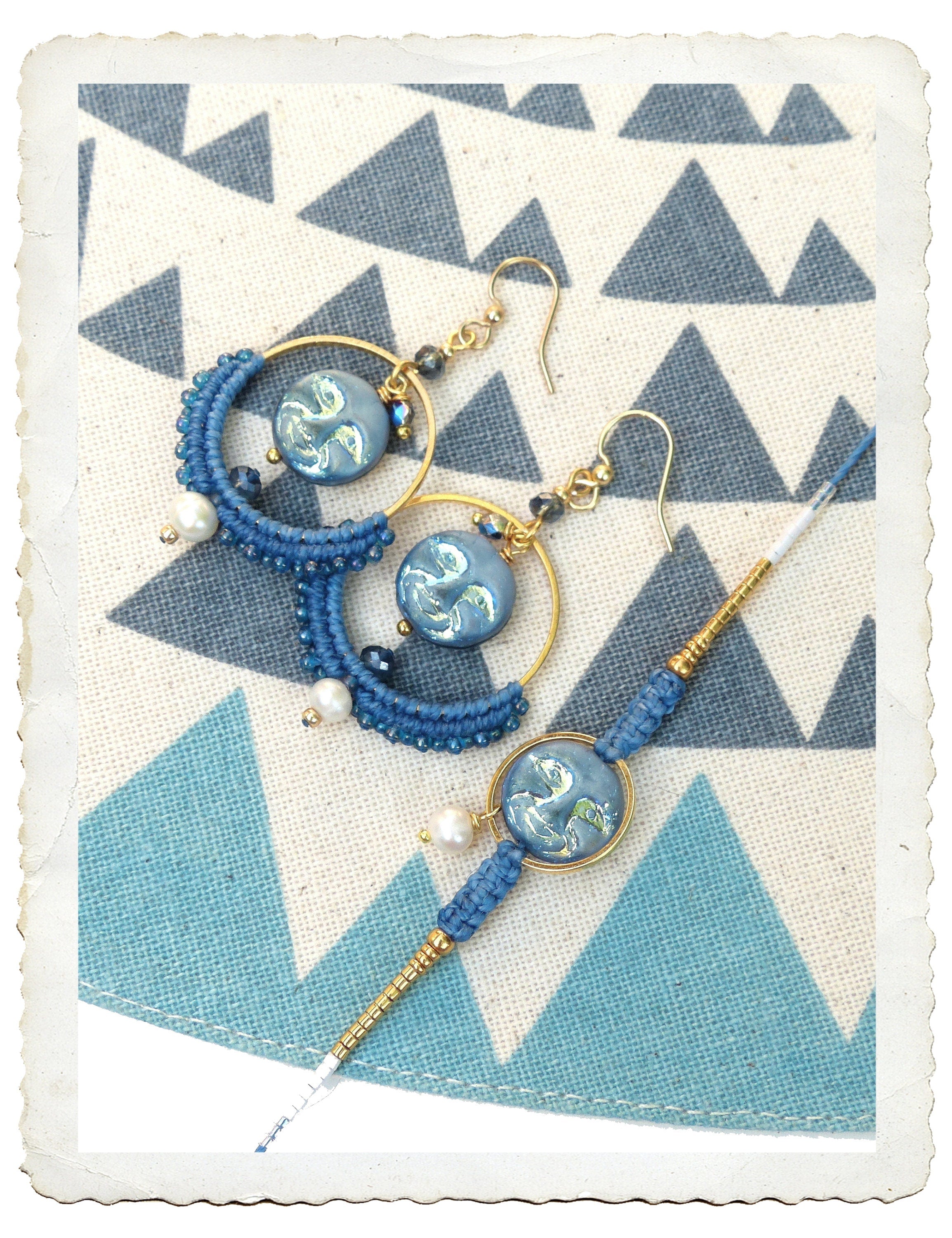 Peut inclure: Une paire de boucles d'oreilles créoles bleues et dorées avec un charme à motif bleu et blanc. Les boucles d'oreilles sont sur un fond de tissu à motif blanc et bleu.