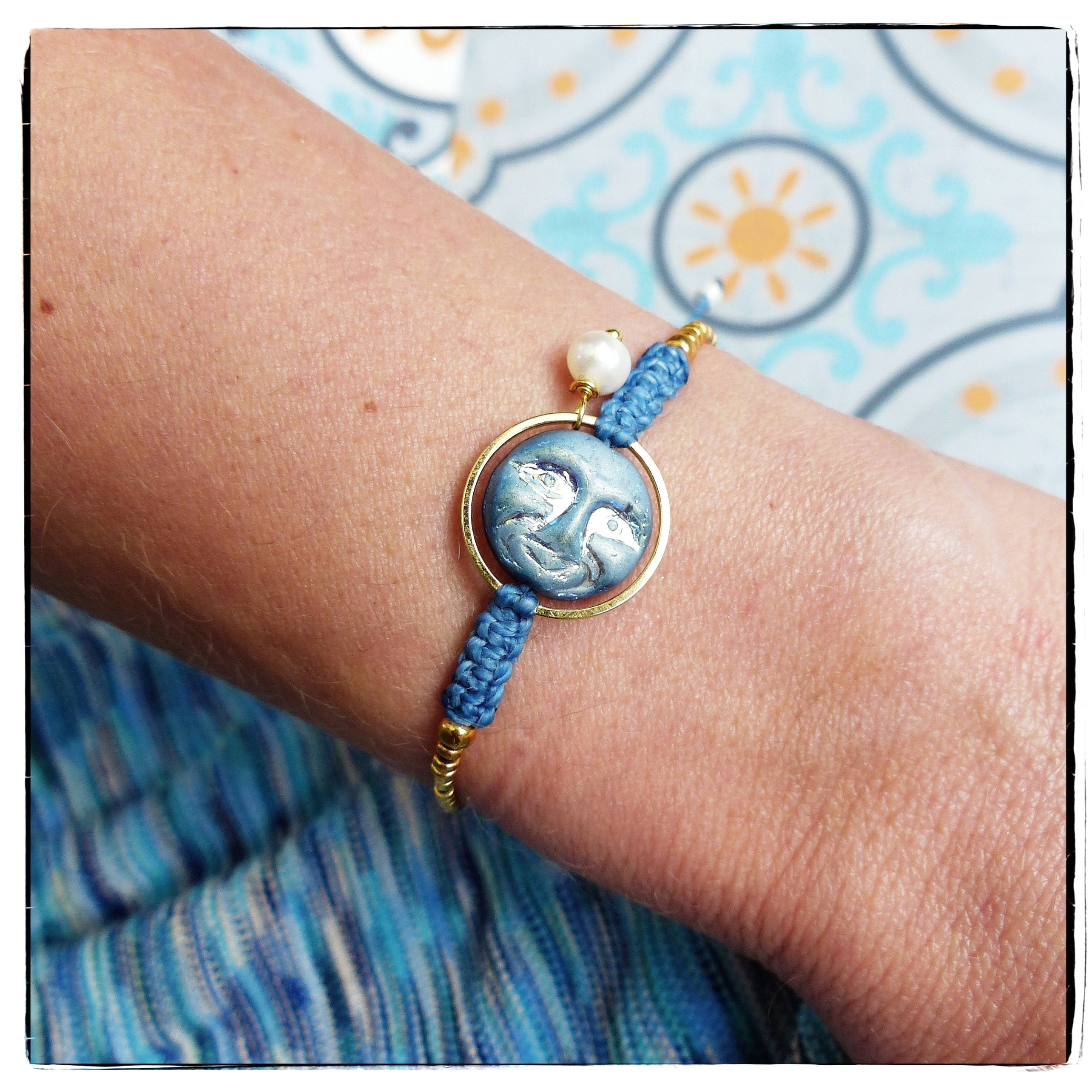 Peut inclure: Un bracelet bleu et or avec un charme visage de lune et un accent de perle blanche. Le bracelet est fait de cordon tressé et a un fermoir en or.