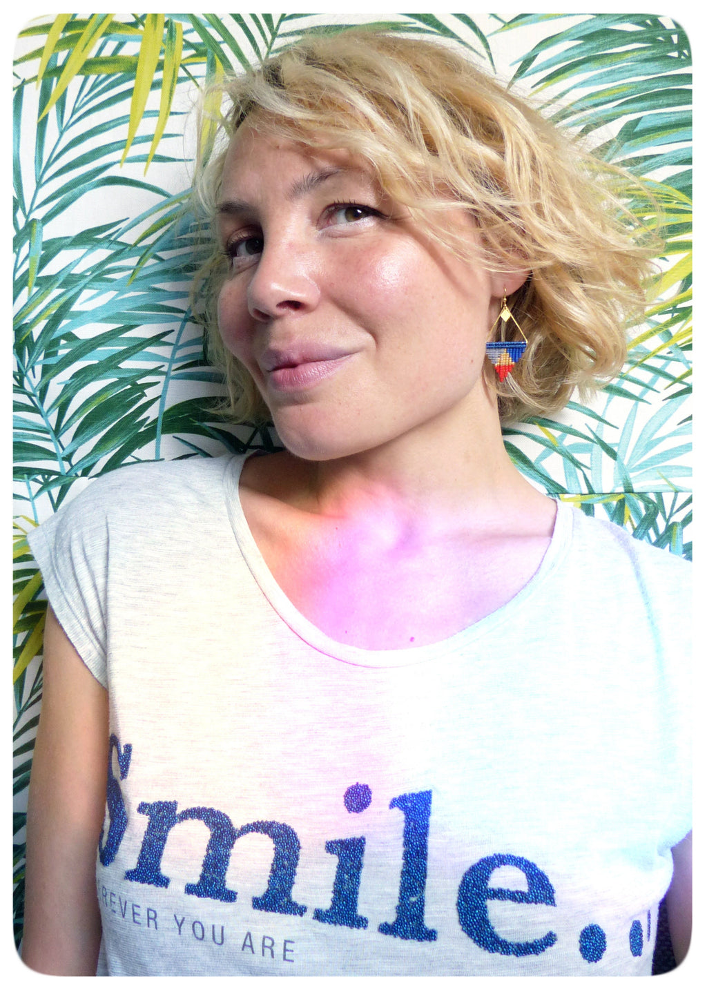 Peut inclure: Une femme aux cheveux blonds porte un t-shirt blanc avec le texte "Smile. Forever You Are" en paillettes bleues. Elle porte une paire de boucles d'oreilles géométriques avec des perles bleues, rouges et jaunes.