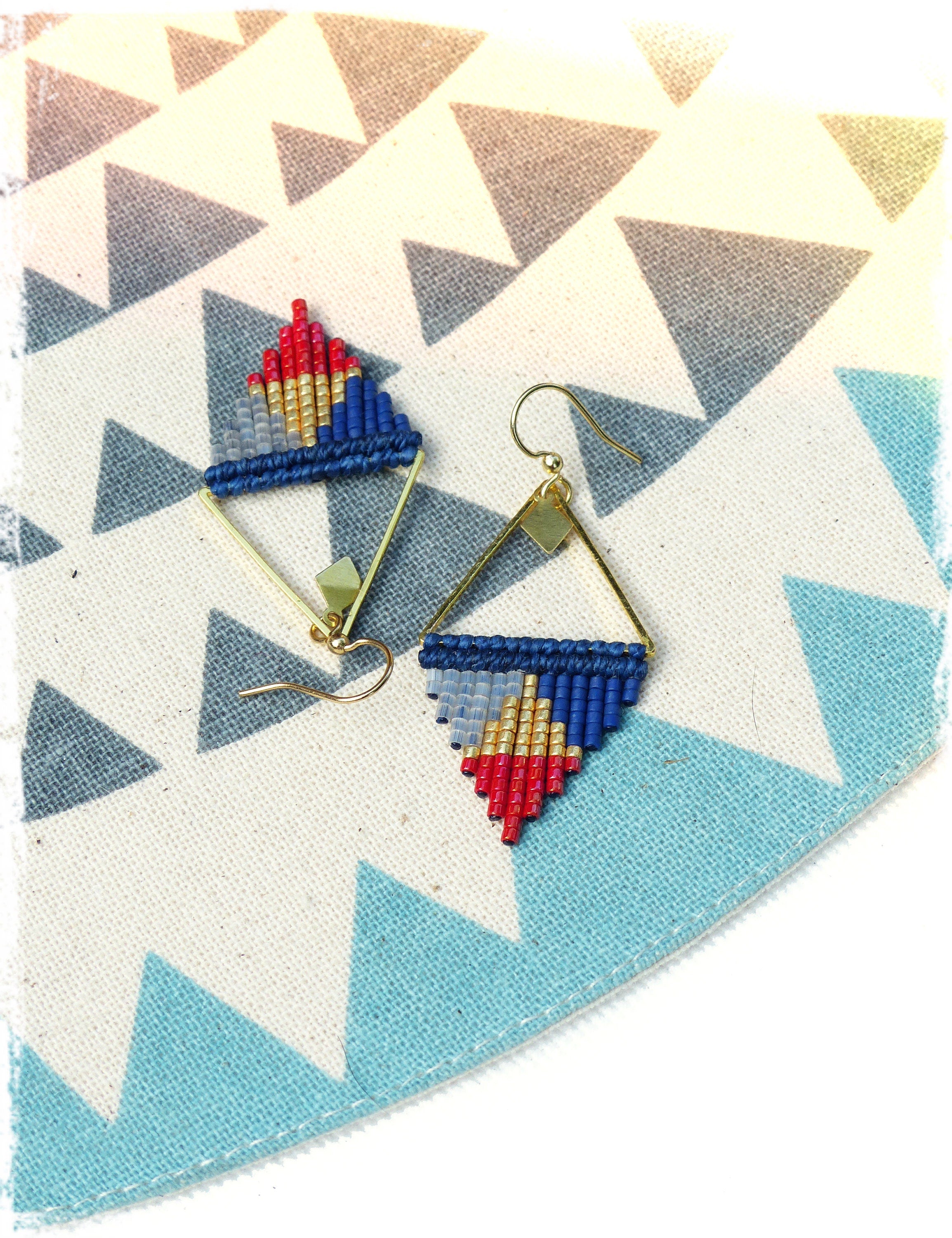 Peut inclure: Une paire de boucles d'oreilles triangulaires dorées avec des franges de perles bleues, rouges et dorées. Les boucles d'oreilles sont sur un fond à motifs blancs et bleus.