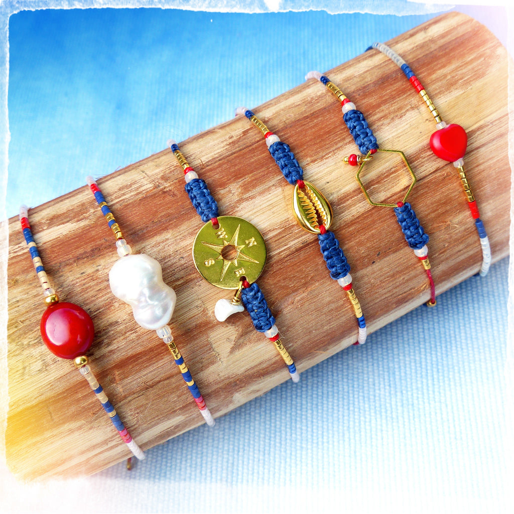 Peut inclure: Six bracelets colorés avec différents pendentifs, dont une boussole, une coquille, un hexagone et un cœur rouge. Les bracelets sont faits de perles bleues, rouges et blanches et sont présentés sur une surface en bois.