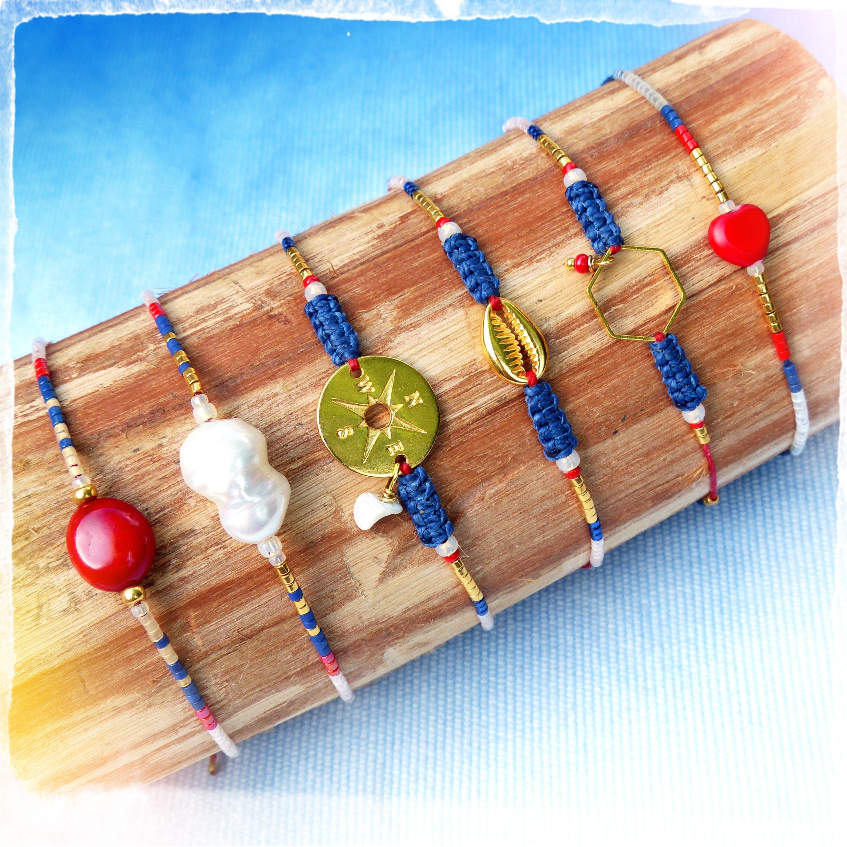 Peut inclure: Six bracelets colorés avec différents pendentifs, dont une boussole, une coquille, un hexagone et un cœur rouge. Les bracelets sont faits de perles bleues, rouges et blanches et sont présentés sur une surface en bois.