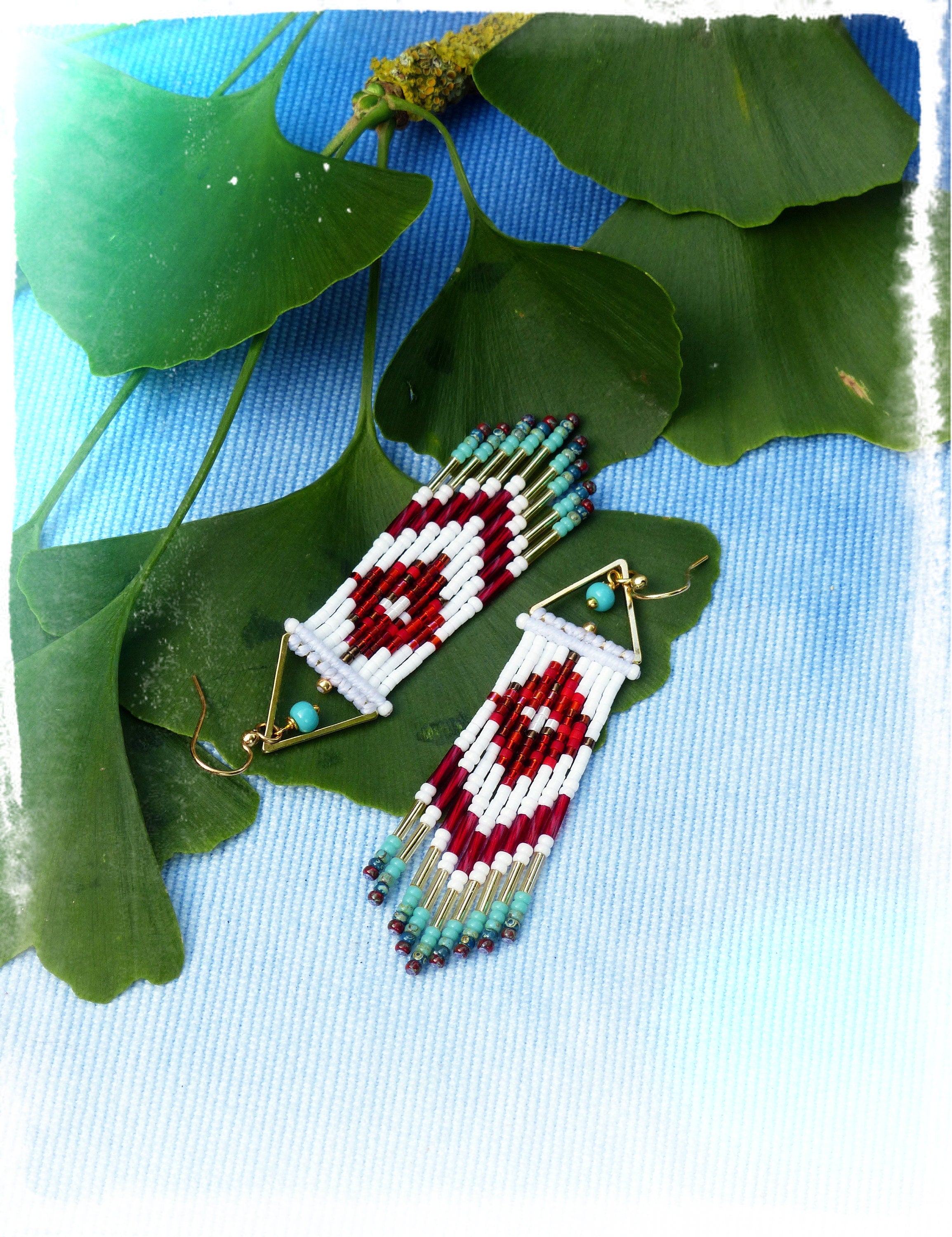 Peut inclure: Une paire de longues boucles d'oreilles en perles avec un motif géométrique. Les boucles d'oreilles sont faites de perles blanches, rouges et turquoise et ont des accents dorés. Les boucles d'oreilles sont suspendues sur un fond bleu.