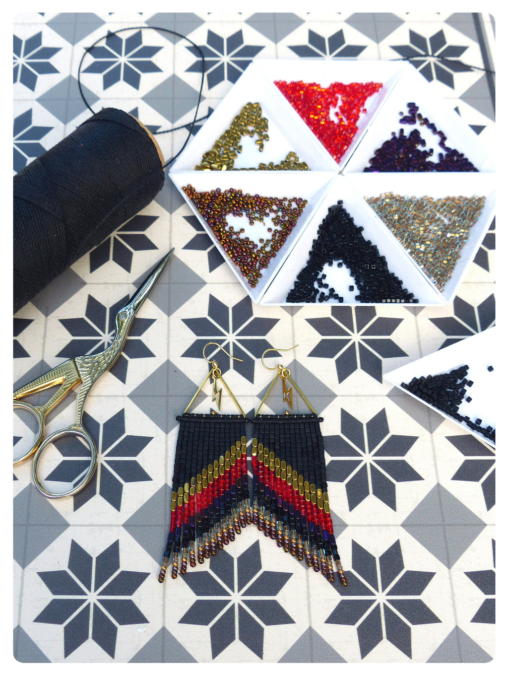 Peut inclure: Boucles d'oreilles faites à la main avec un motif en chevrons noir, rouge et or. Les boucles d'oreilles ont un dessus triangulaire avec un charme éclair doré. L'image montre également une bobine de fil noir, des ciseaux et des petits plateaux de perles.