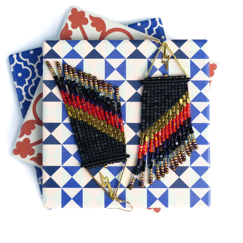 Peut inclure: Paire de boucles d'oreilles perlées faites à la main, noires, rouges, dorées et bleues. Les boucles d'oreilles présentent un motif géométrique avec un éclair et une frange. Présentées sur une surface à motifs.