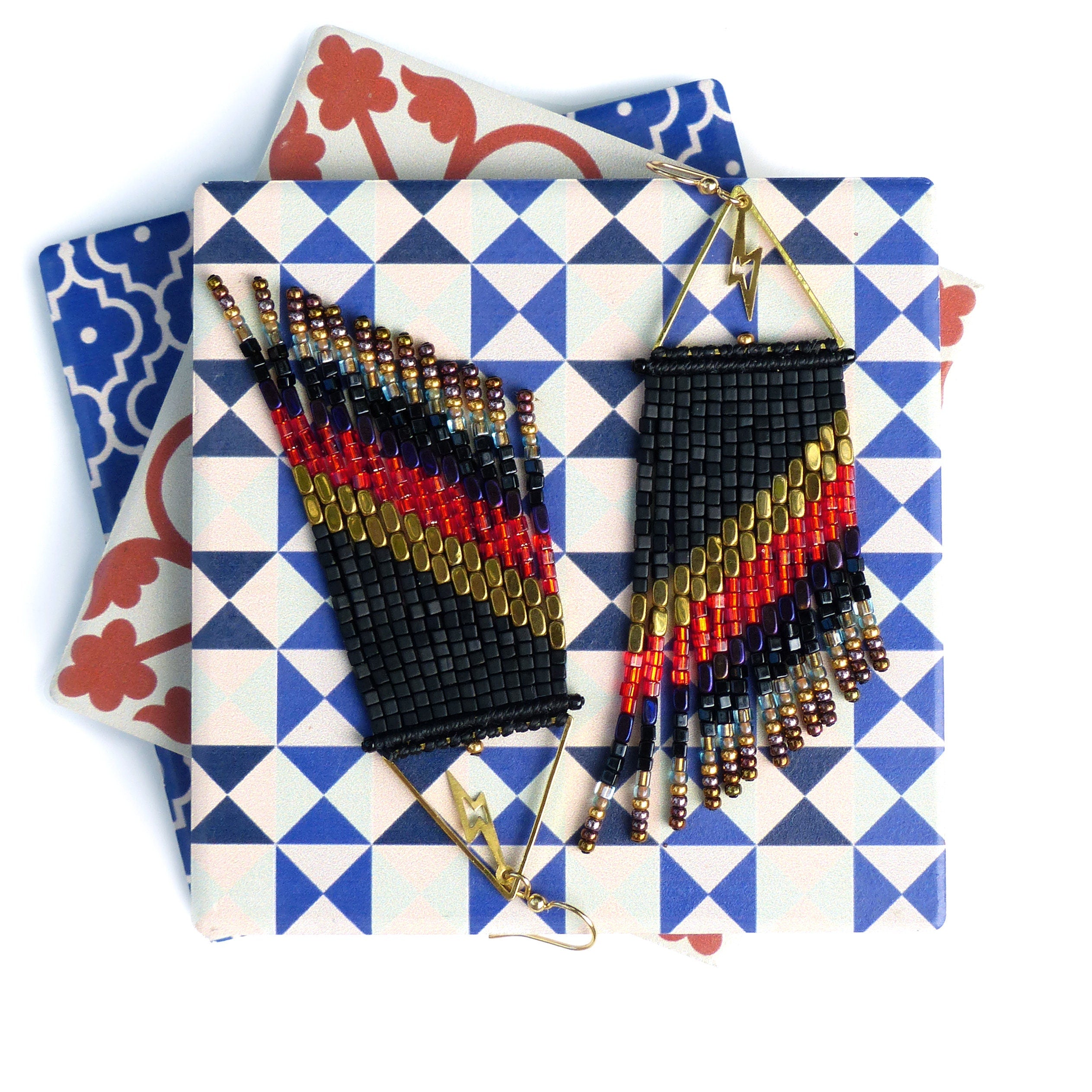 Peut inclure: Paire de boucles d'oreilles perlées faites à la main, noires, rouges, dorées et bleues. Les boucles d'oreilles présentent un motif géométrique avec un éclair et une frange. Présentées sur une surface à motifs.