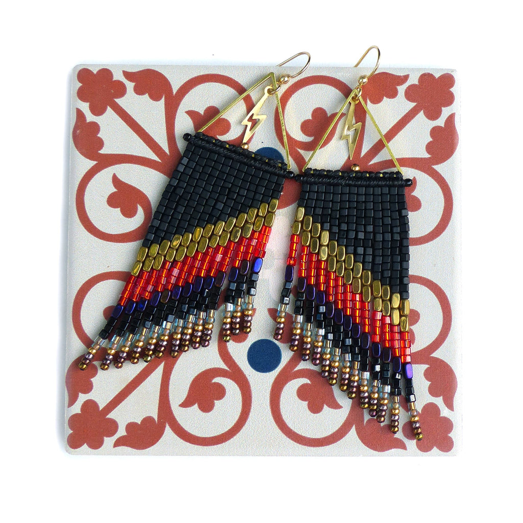 Peut inclure: Boucles d'oreilles en perles avec un dégradé noir, or, rouge et violet. Les boucles d'oreilles ont des franges et des éclairs dorés. Elles sont présentées sur un carreau décoratif.