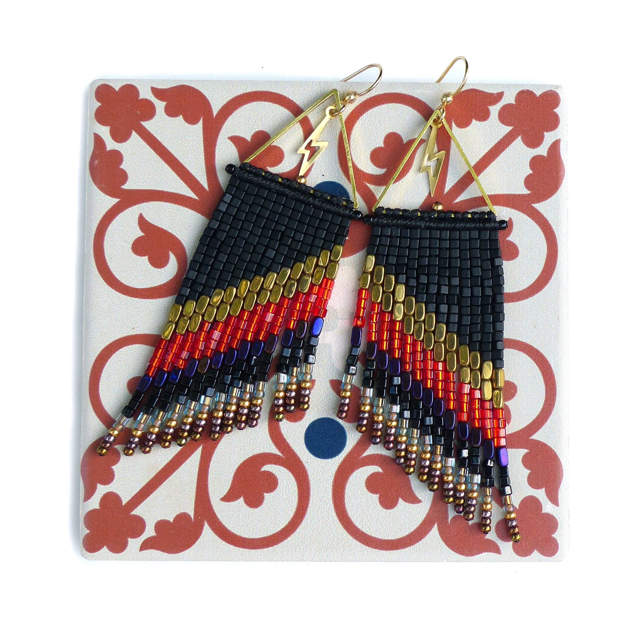 Peut inclure: Boucles d'oreilles en perles avec un dégradé noir, or, rouge et violet. Les boucles d'oreilles ont des franges et des éclairs dorés. Elles sont présentées sur un carreau décoratif.