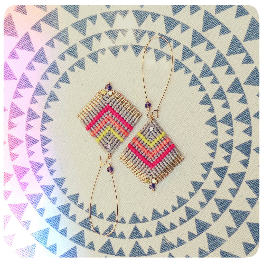 Peut inclure: Une paire de boucles d'oreilles à motifs géométriques avec des anneaux dorés et du fil rose, jaune et gris. Les boucles d'oreilles sont sur un fond blanc avec un motif de triangles gris.