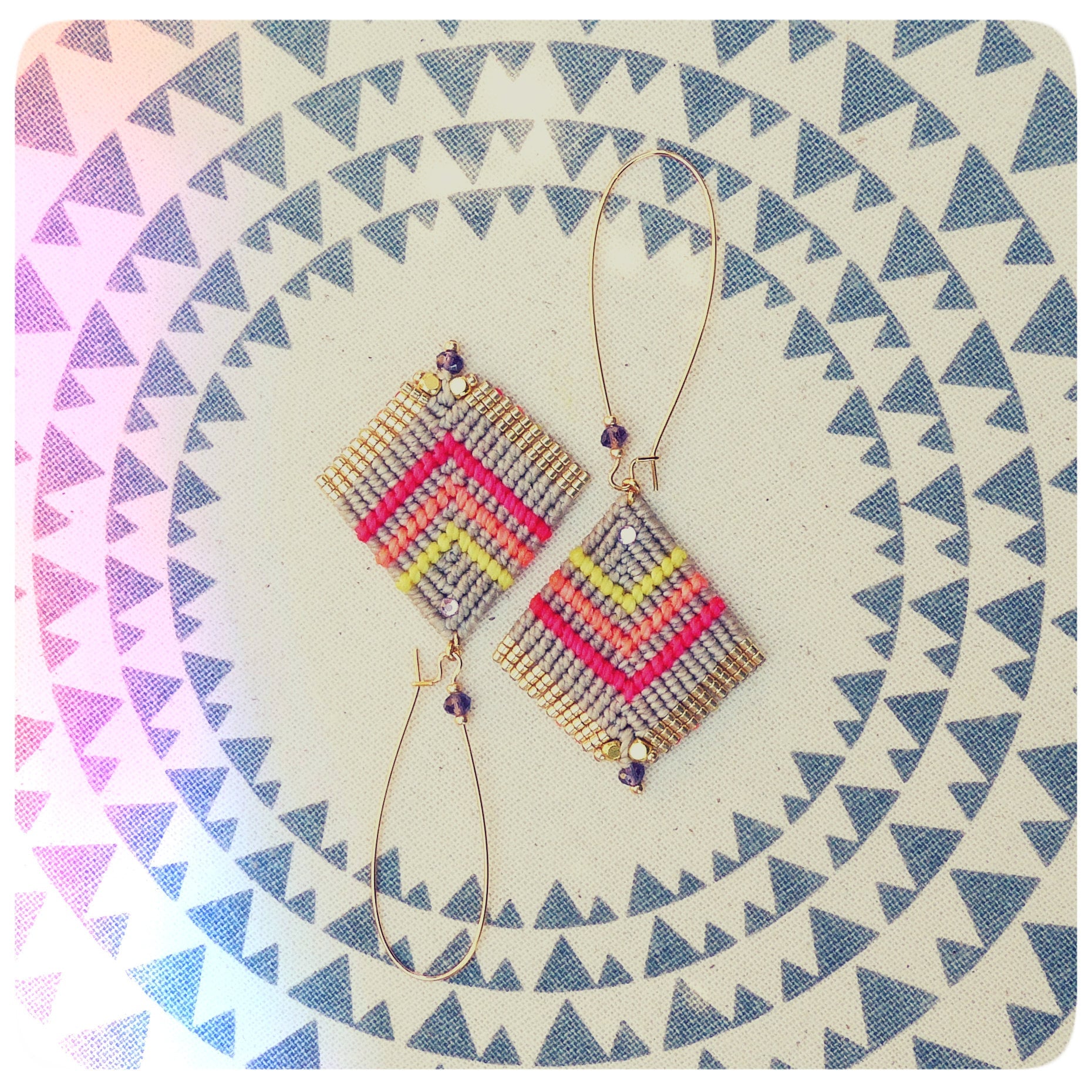 Peut inclure: Une paire de boucles d'oreilles à motifs géométriques avec des anneaux dorés et du fil rose, jaune et gris. Les boucles d'oreilles sont sur un fond blanc avec un motif de triangles gris.