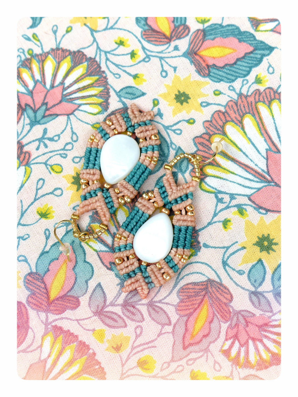 Peut inclure: Une paire de boucles d'oreilles en macramé faites à la main avec une pierre blanche en forme de larme et un cordon de couleur pêche. Les boucles d'oreilles sont sur un fond de tissu floral.