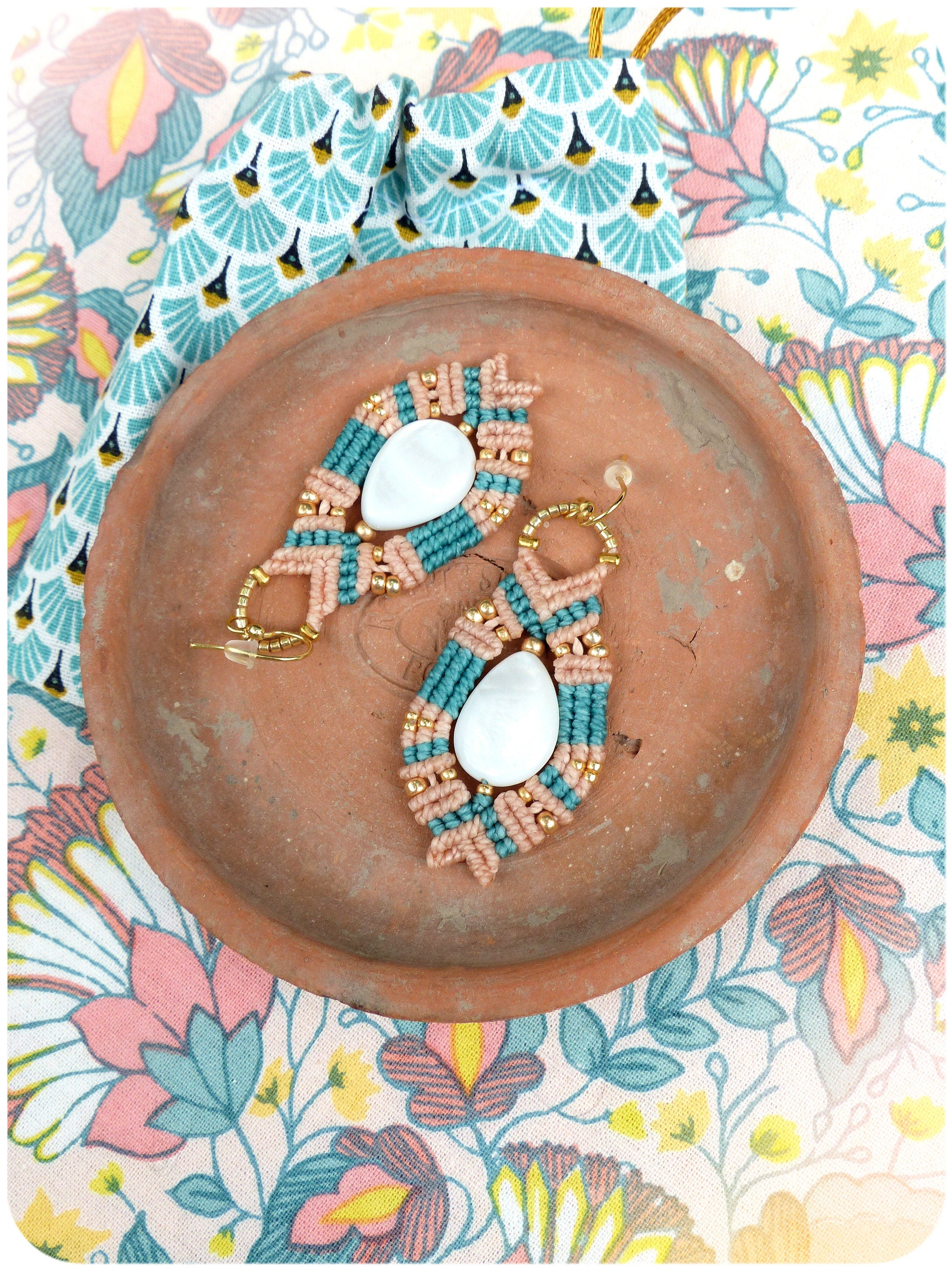 Peut inclure: Une paire de boucles d'oreilles faites à la main avec un pendentif en coquille blanche, du macramé rose et turquoise et des accents dorés. Les boucles d'oreilles sont présentées sur une assiette en terre cuite à motif floral.