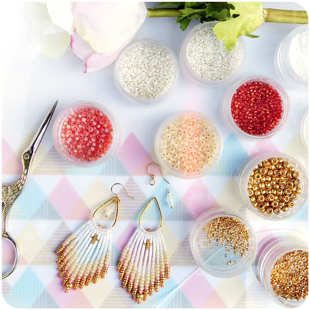 Peut inclure: Une paire de boucles d'oreilles à franges en perles blanches et dorées. Les boucles d'oreilles sont présentées sur une surface blanche avec de petits contenants de perles de différentes couleurs.