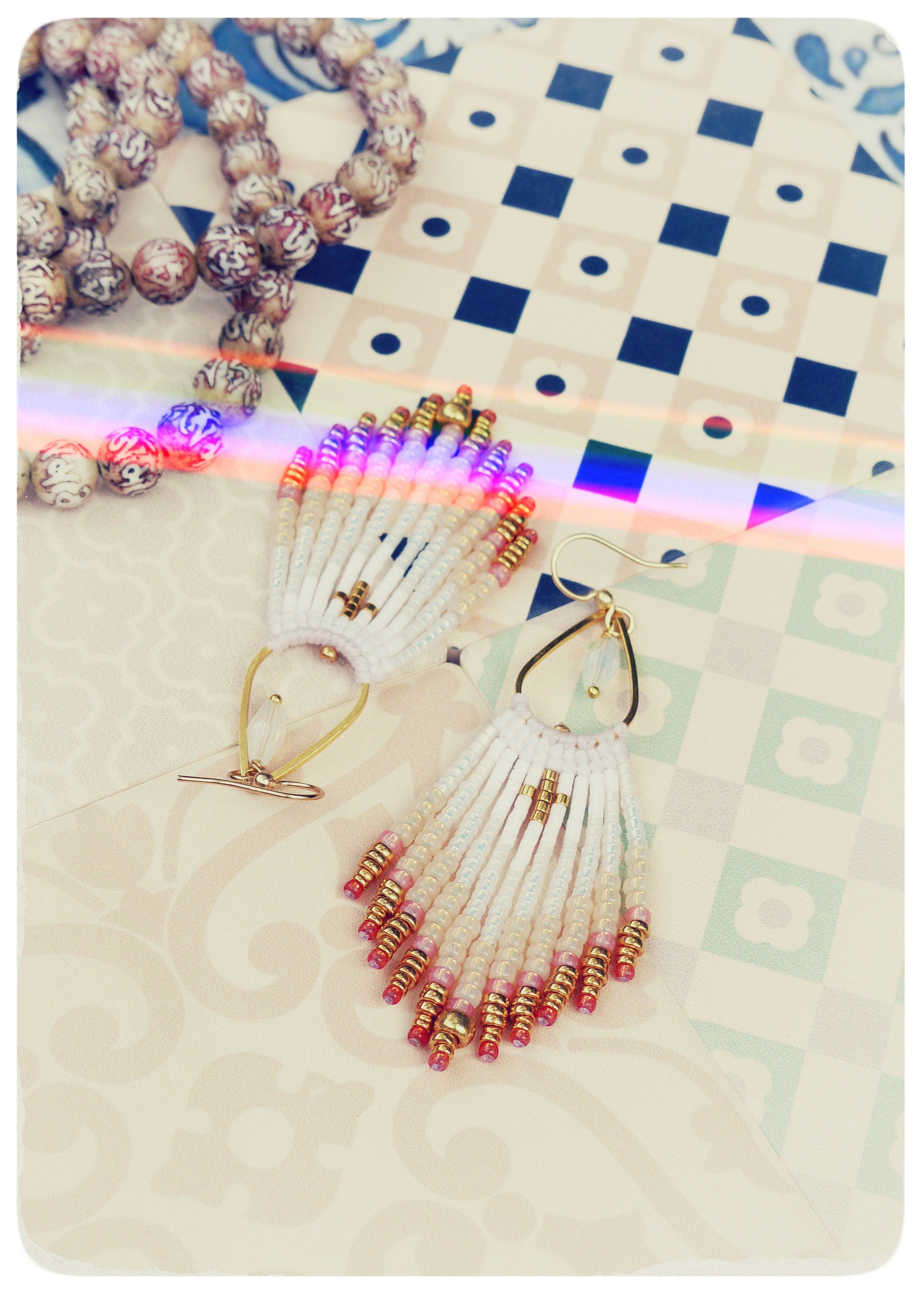 Peut inclure: Une paire de boucles d'oreilles en perles blanches et dorées avec des accents roses et dorés. Les boucles d'oreilles sont pendantes et ont un design en forme de glands.