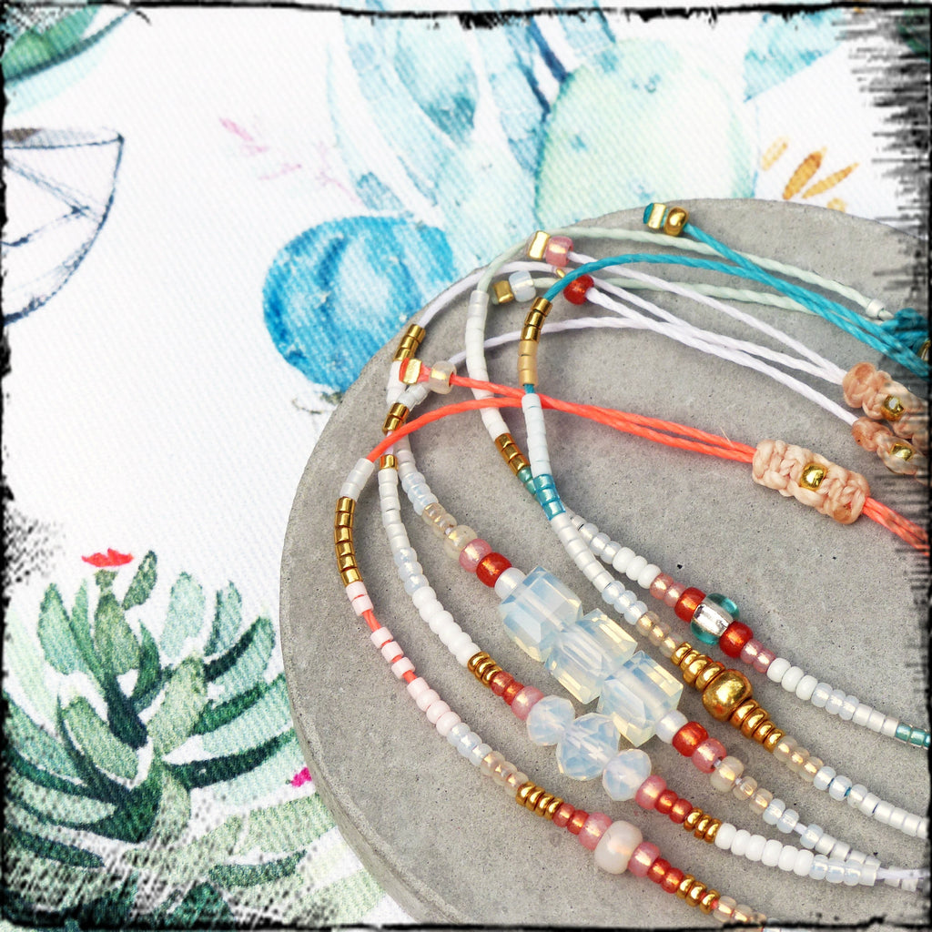 Peut inclure: Un ensemble de bracelets en perles colorées avec des perles blanches, roses, bleues et dorées. Les bracelets sont superposés sur une surface grise.