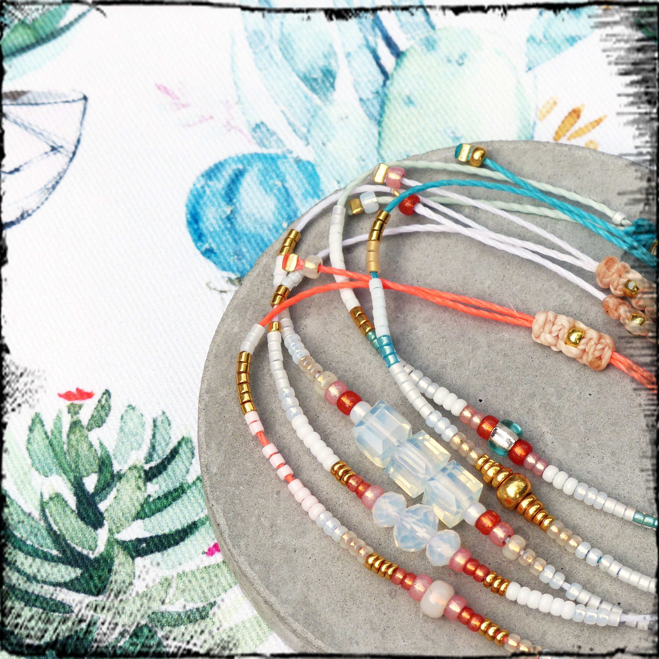Peut inclure: Un ensemble de bracelets en perles colorées avec des perles blanches, roses, bleues et dorées. Les bracelets sont superposés sur une surface grise.