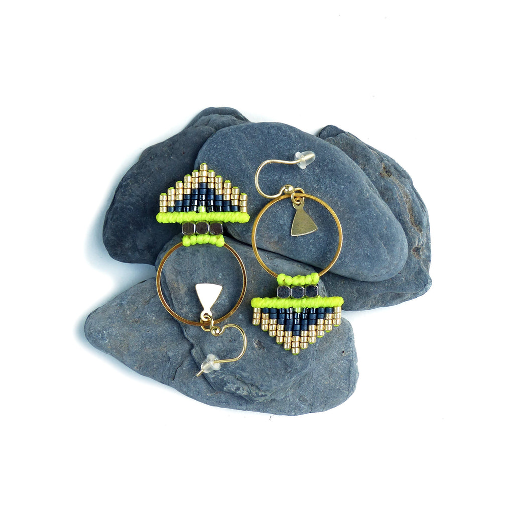 Peut inclure: Une paire de boucles d'oreilles créoles dorées avec des motifs géométriques en perles noires et vertes. Les boucles d'oreilles sont présentées sur deux pierres grises.