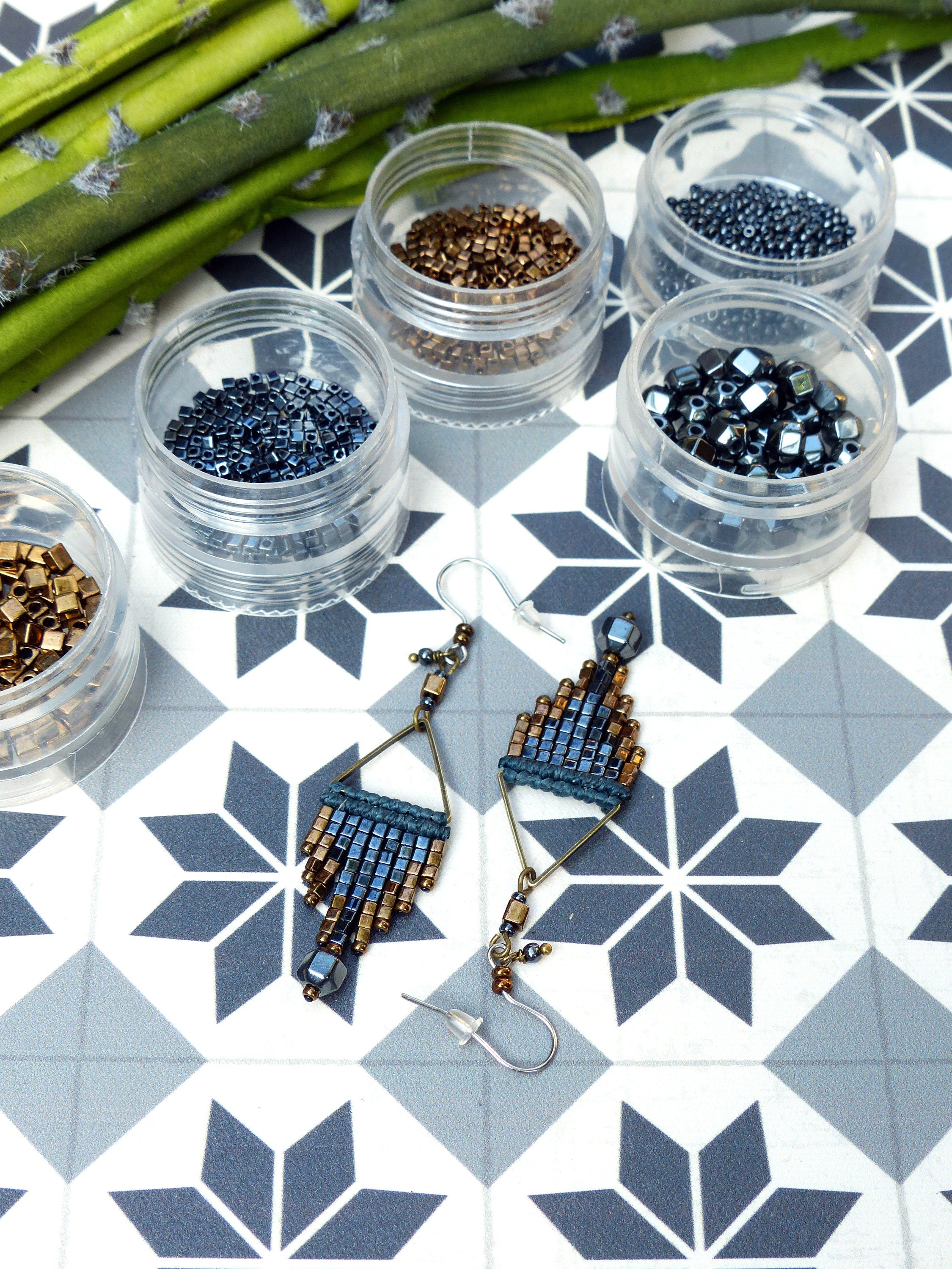 Peut inclure: Une paire de boucles d'oreilles faites à la main avec un design géométrique. Les boucles d'oreilles sont faites de perles bleues et dorées et ont une forme triangulaire. Les boucles d'oreilles sont suspendues à des crochets argentés.